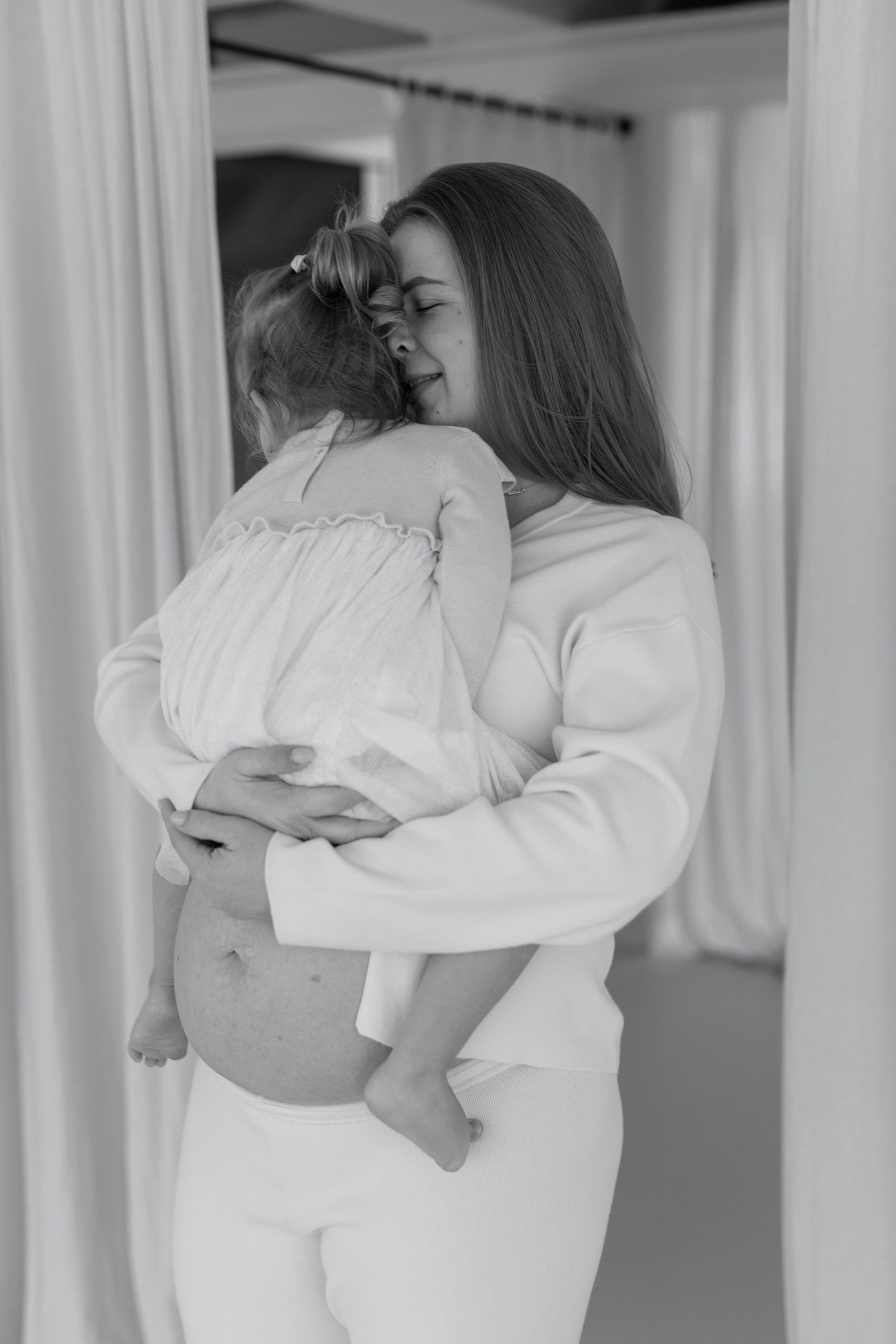 Black and white photo of a pregnant woman in white holding and embracing a young child indoors.