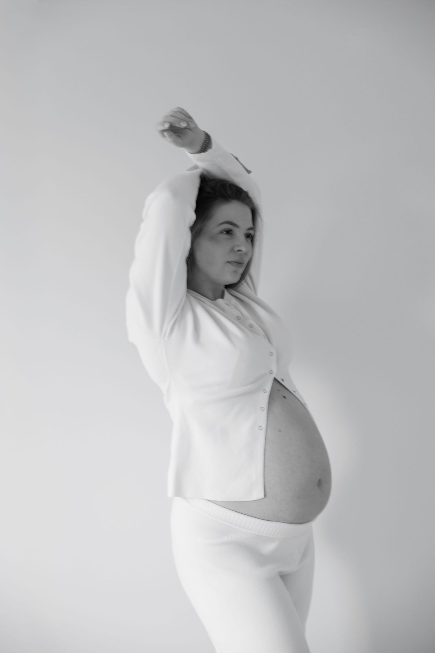 Black and white photo of a pregnant woman in white outfit posing with arms raised, showcasing her baby bump.