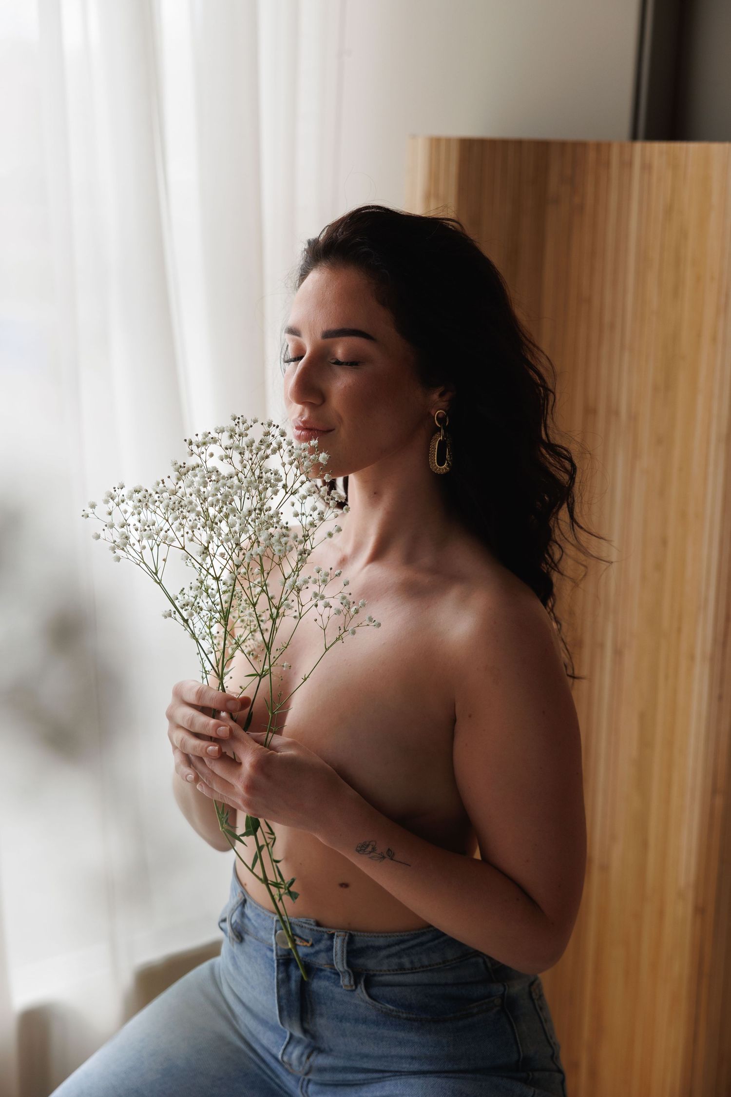 Woman with dark hair holding white baby's breath flowers, wearing jeans, leaning against wooden wall near window.