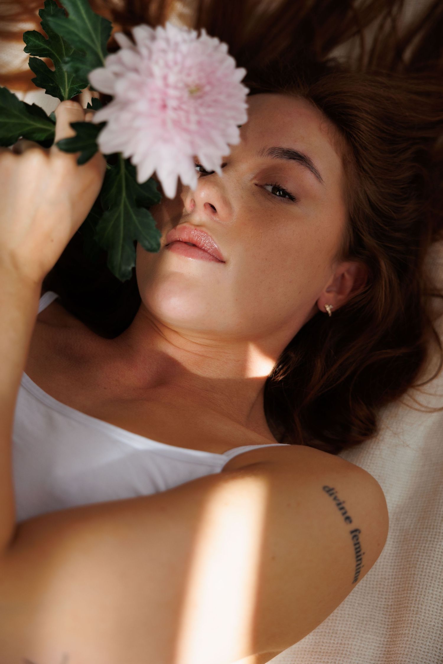 Young woman lying down holding a pink chrysanthemum flower near her face, with a tattoo on her arm.