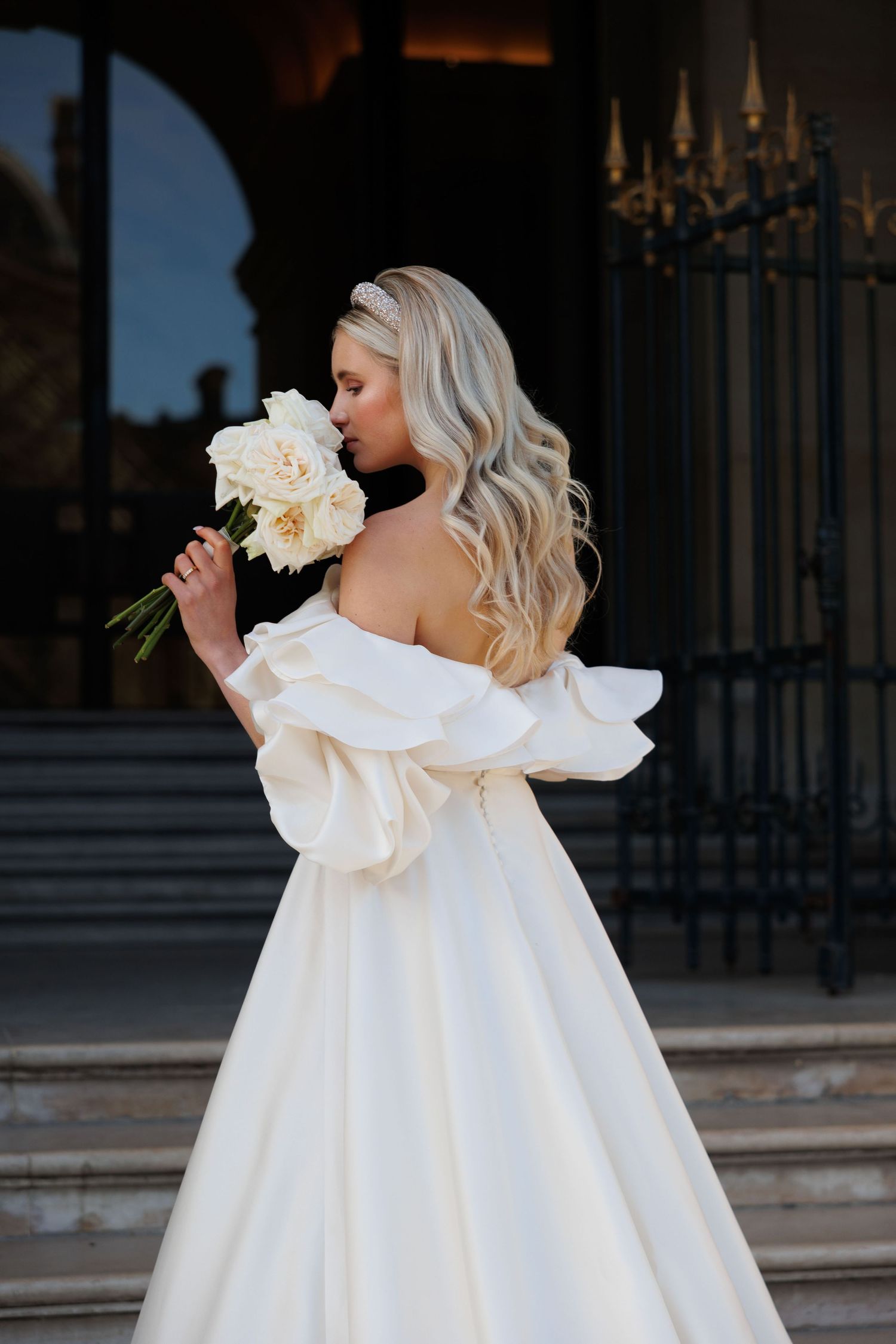 A bride in an elegant off-shoulder white gown smells a cream-colored peony bouquet against an ornate building backdrop.