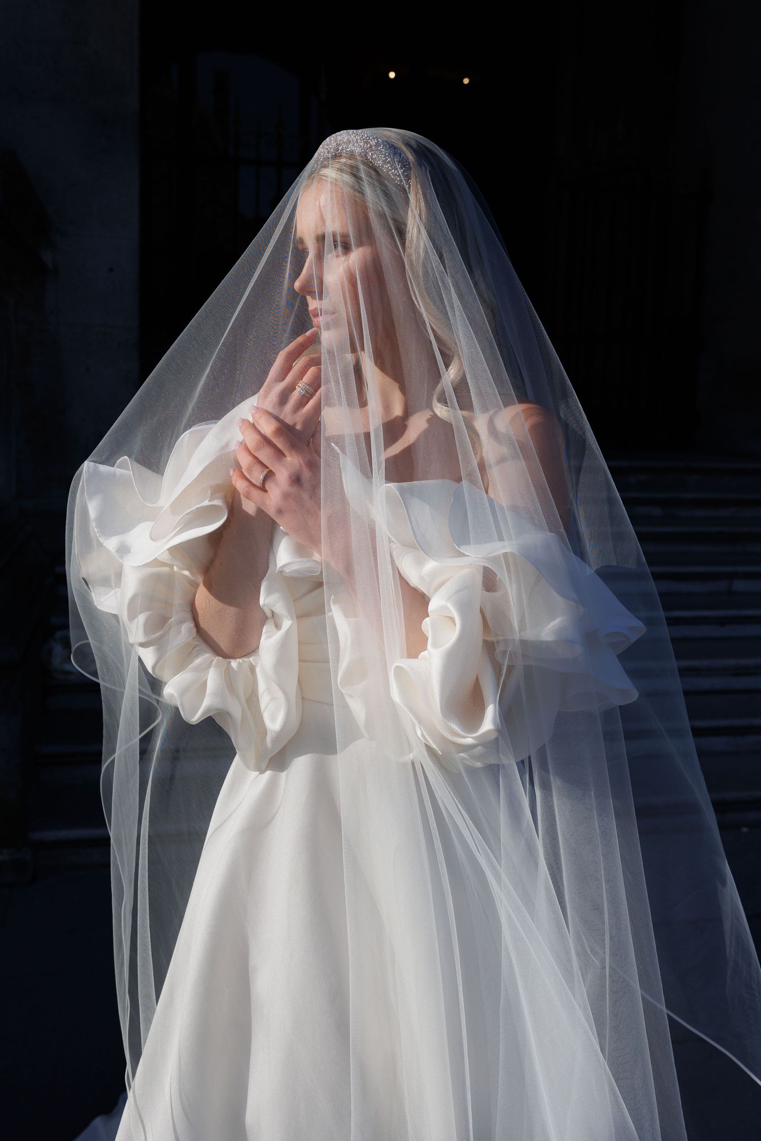 Elegant bride in a white off-shoulder gown with ruffled sleeves, draped in a sheer tulle veil, posing gracefully.