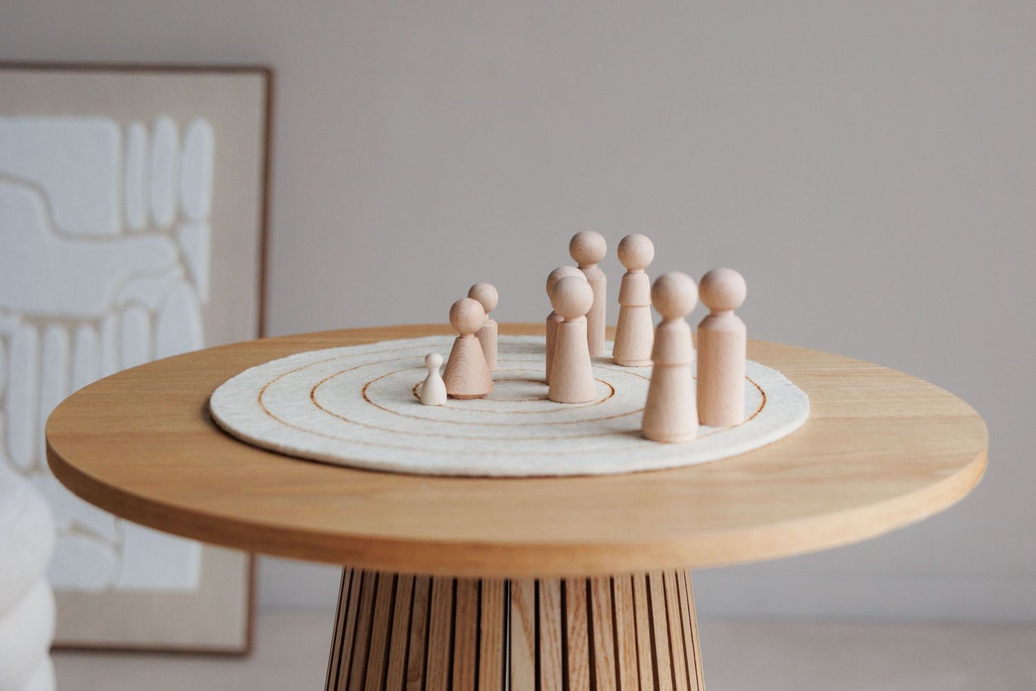Wooden peg dolls arranged on a circular layered wooden board atop a round natural wood table in a minimalist room.
