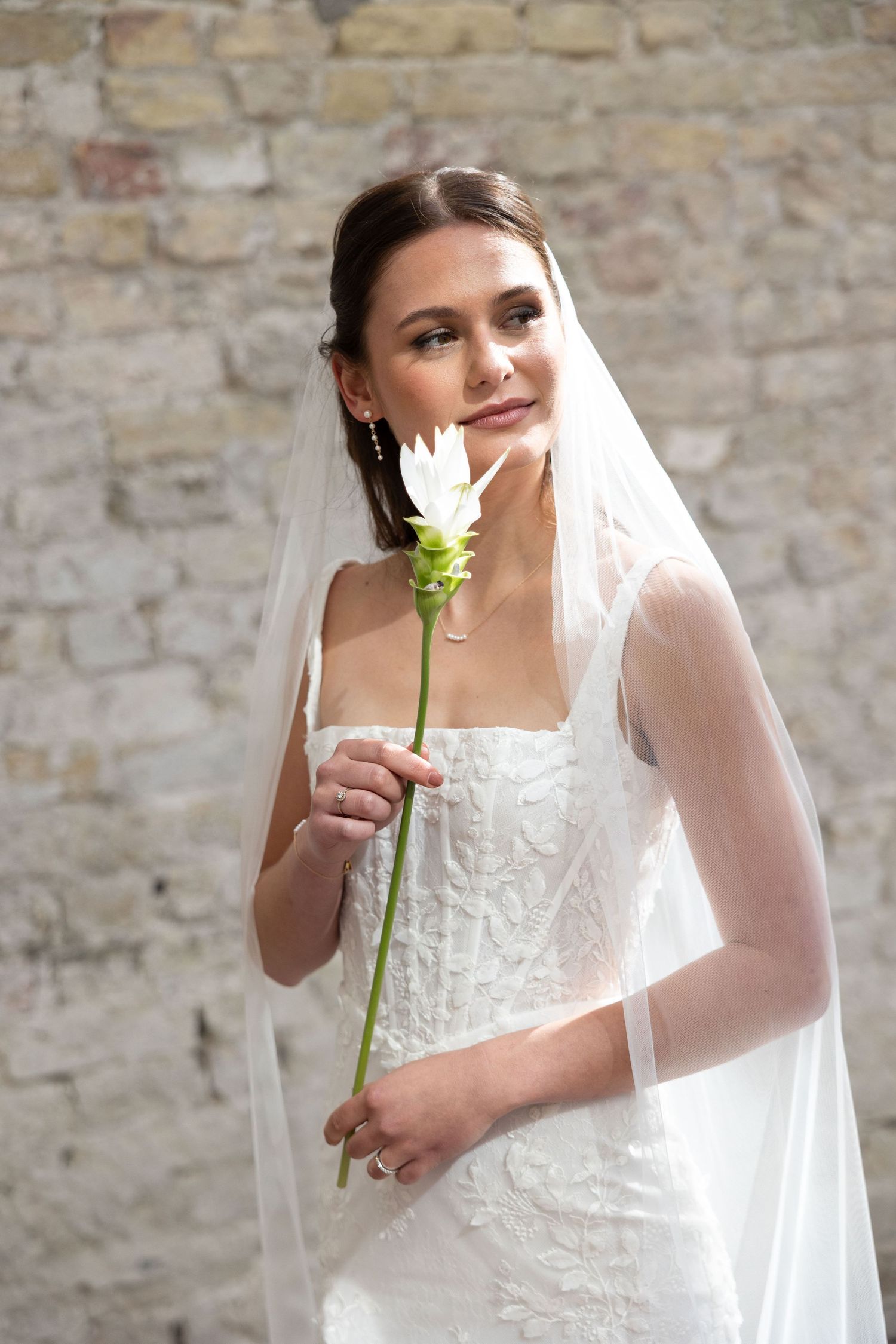 Bride in white lace gown and veil holds a single white flower, posing gracefully against a stone wall backdrop.