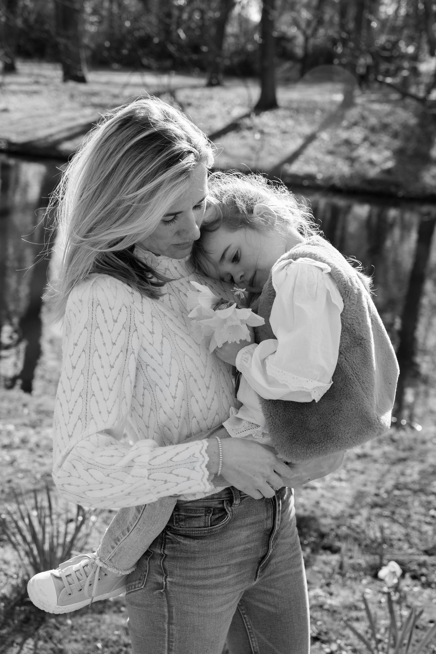 Black and white photo of a mother tenderly holding her young daughter outdoors near a pond.