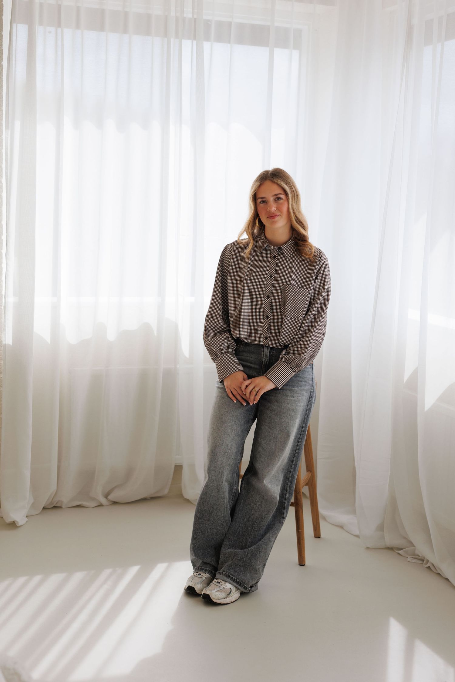 Young woman in gray wide-leg jeans and striped shirt standing near white curtains in bright room.
