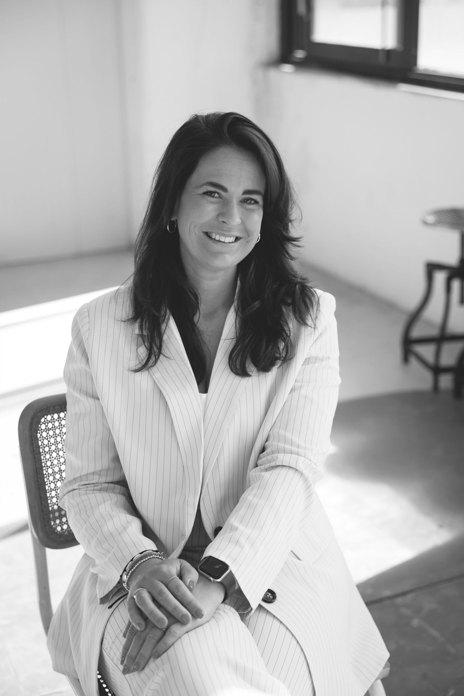 Black and white portrait of a smiling woman in a striped blazer seated on a chair in a bright modern room.