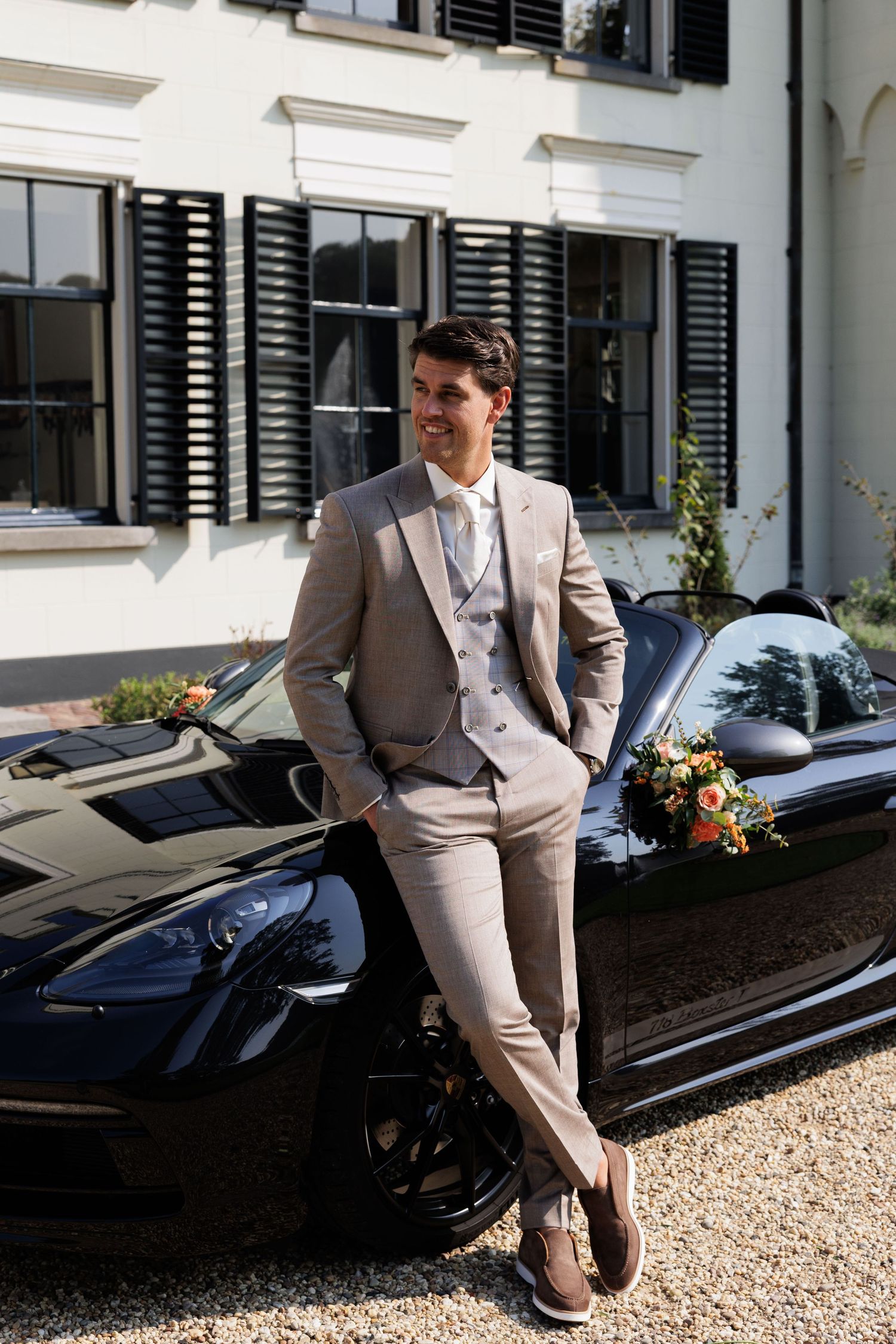 Groom in beige suit leaning against black Porsche with floral bouquet outside white mansion.