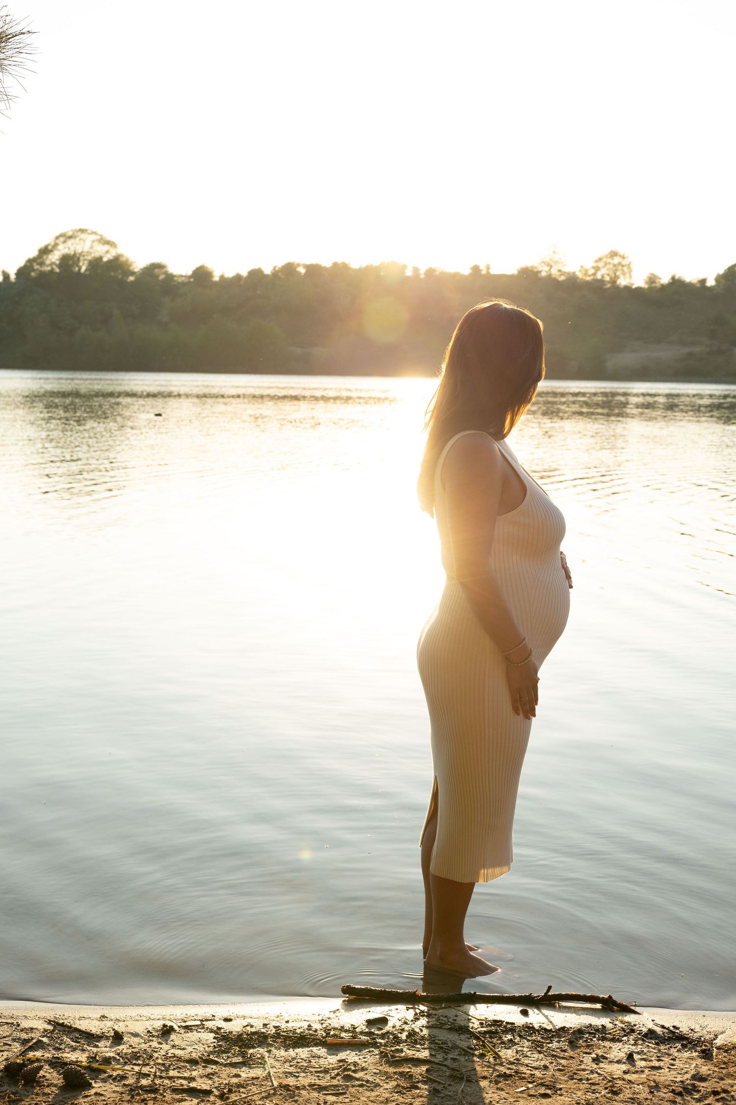 Pregnant woman in beige dress standing barefoot by a sunlit lake at golden hour, cradling her baby bump.