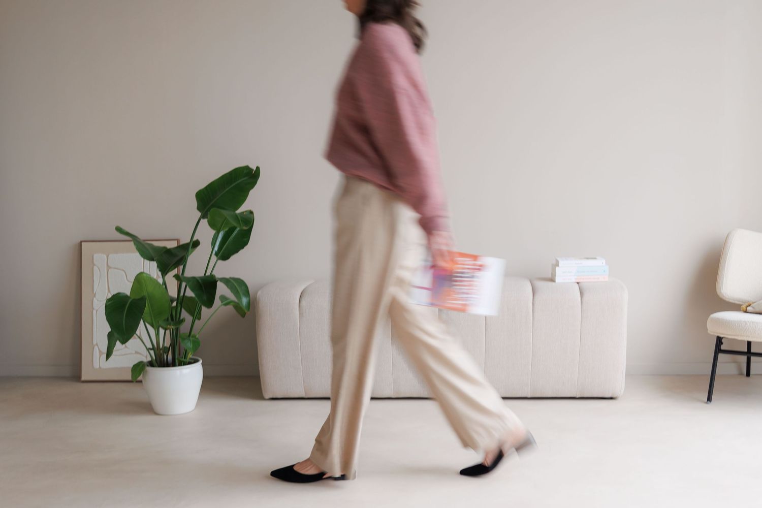 Blurred motion of a woman in pink sweater and beige pants walking through a minimalist white room with a plant.