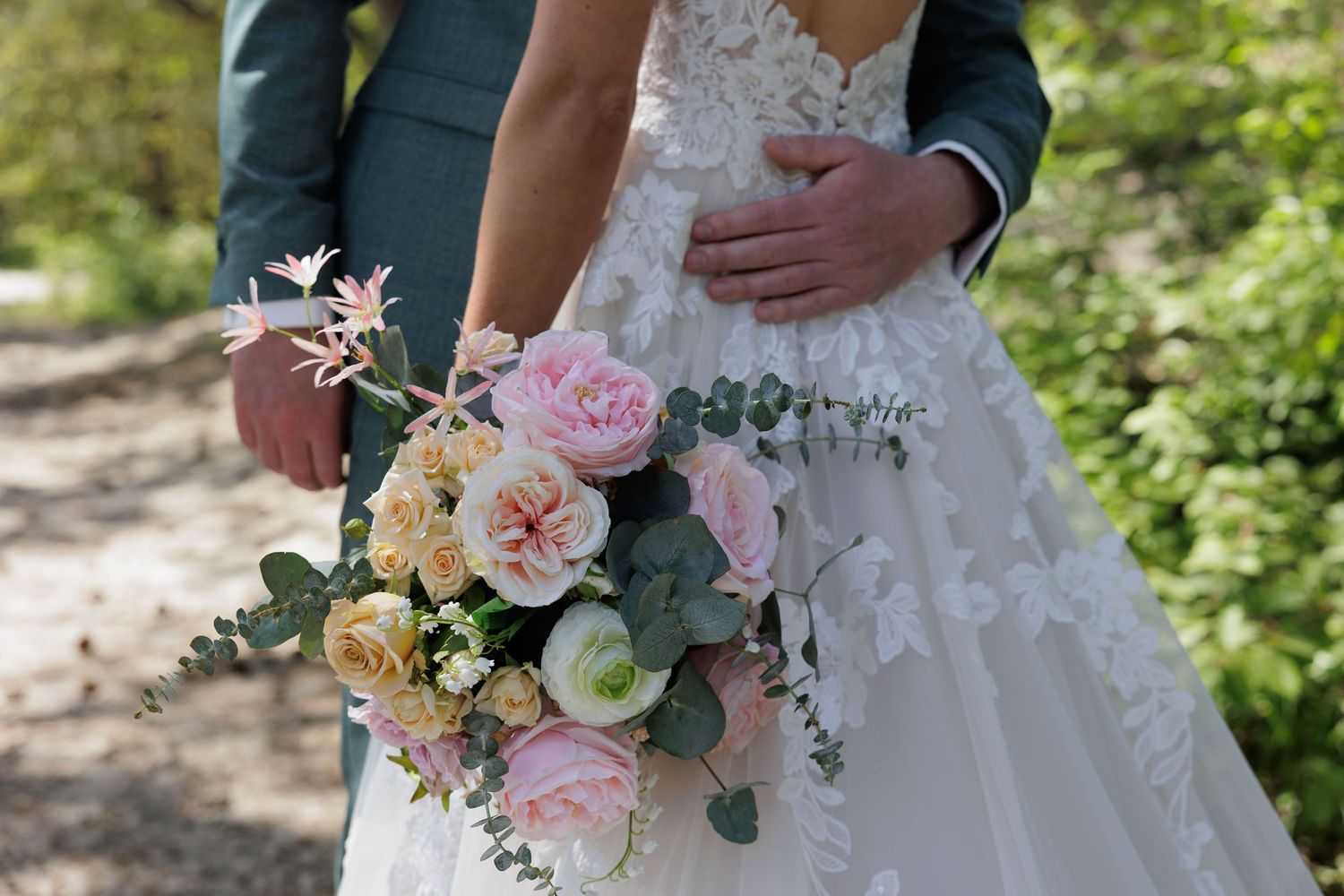 Bride in lace wedding dress holding a cascading bouquet of pink and cream roses with eucalyptus, groom in teal suit.