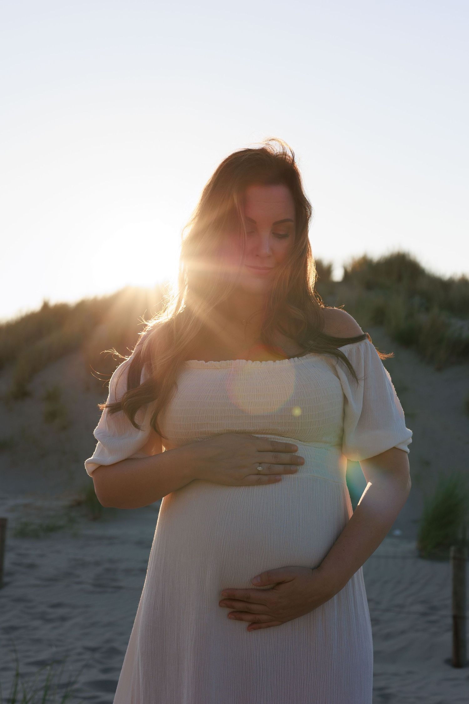 Pregnant woman in white dress cradling her baby bump on a beach at golden sunset.