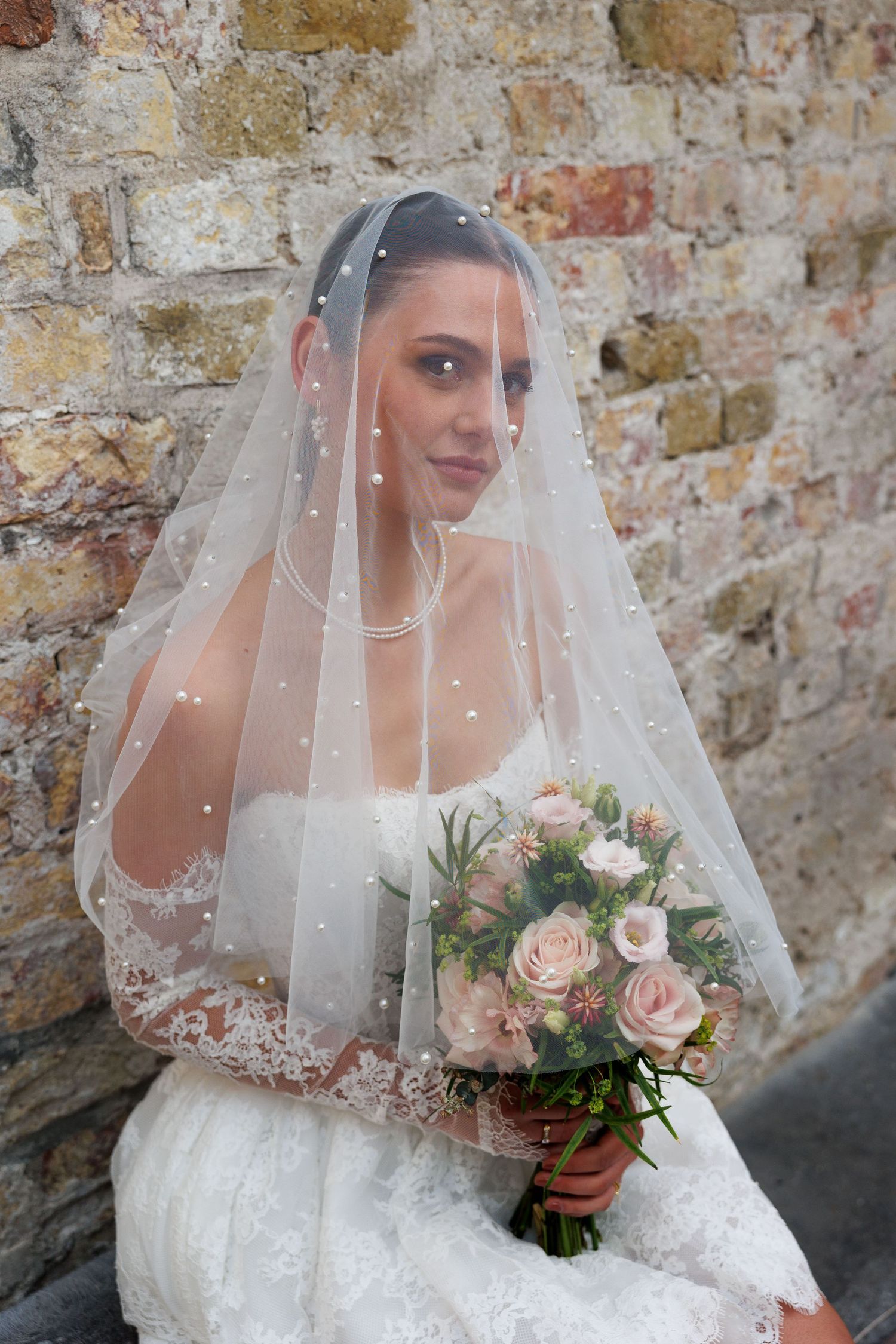 Bride wearing a pearl-adorned veil holding a pink rose bouquet, posed against a rustic stone wall in a lace wedding dress.