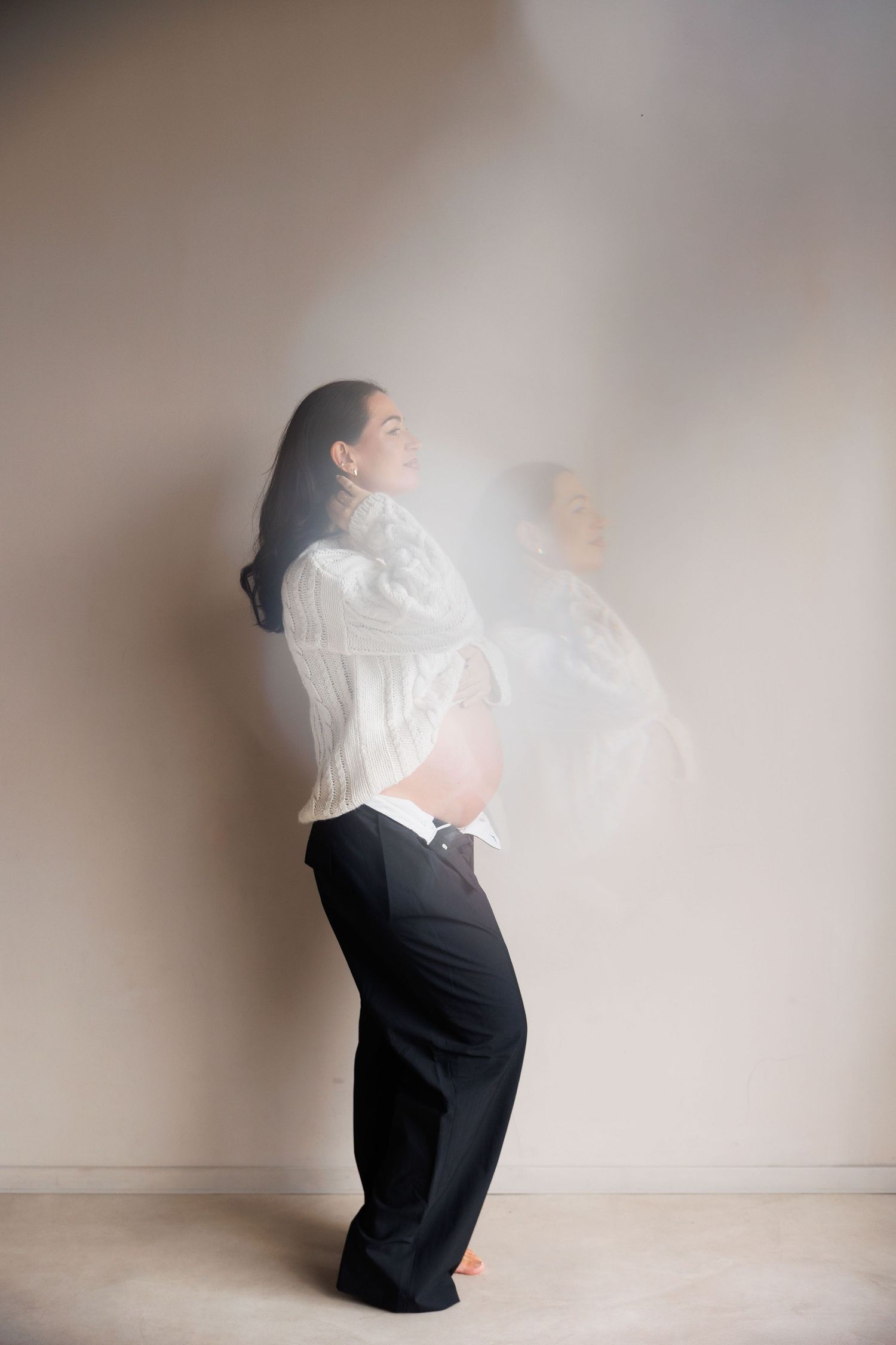 Pregnant woman in white blouse and black trousers poses gracefully against a neutral wall with soft motion blur effect.