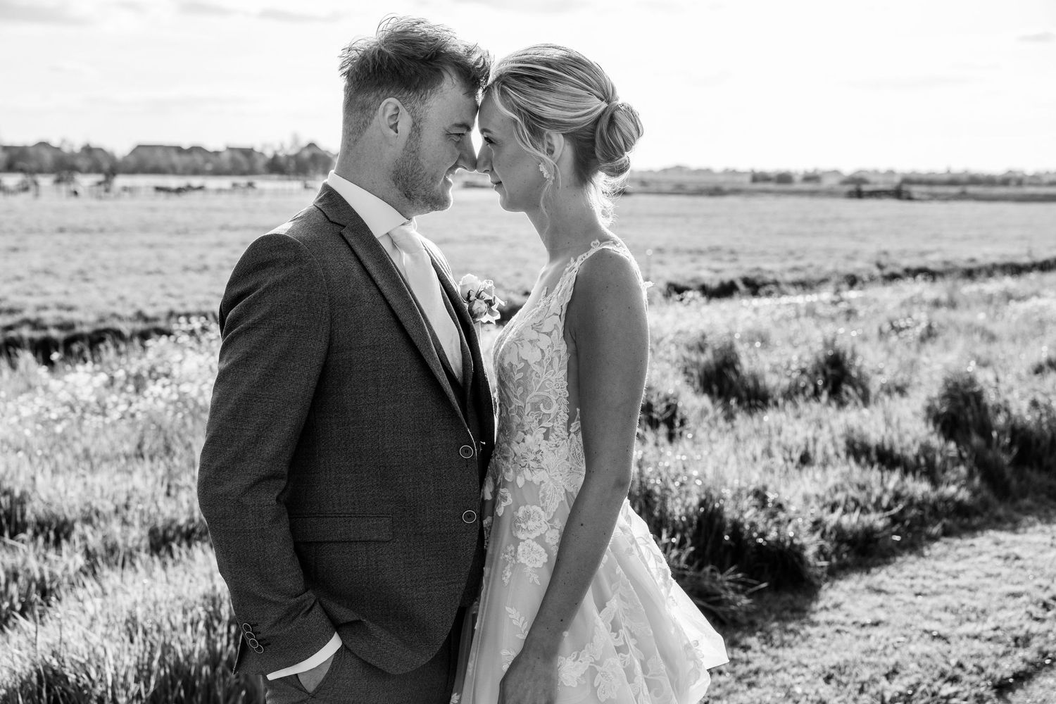 Black and white wedding photo of a couple touching foreheads in a field, bride in lace gown and groom in suit.
