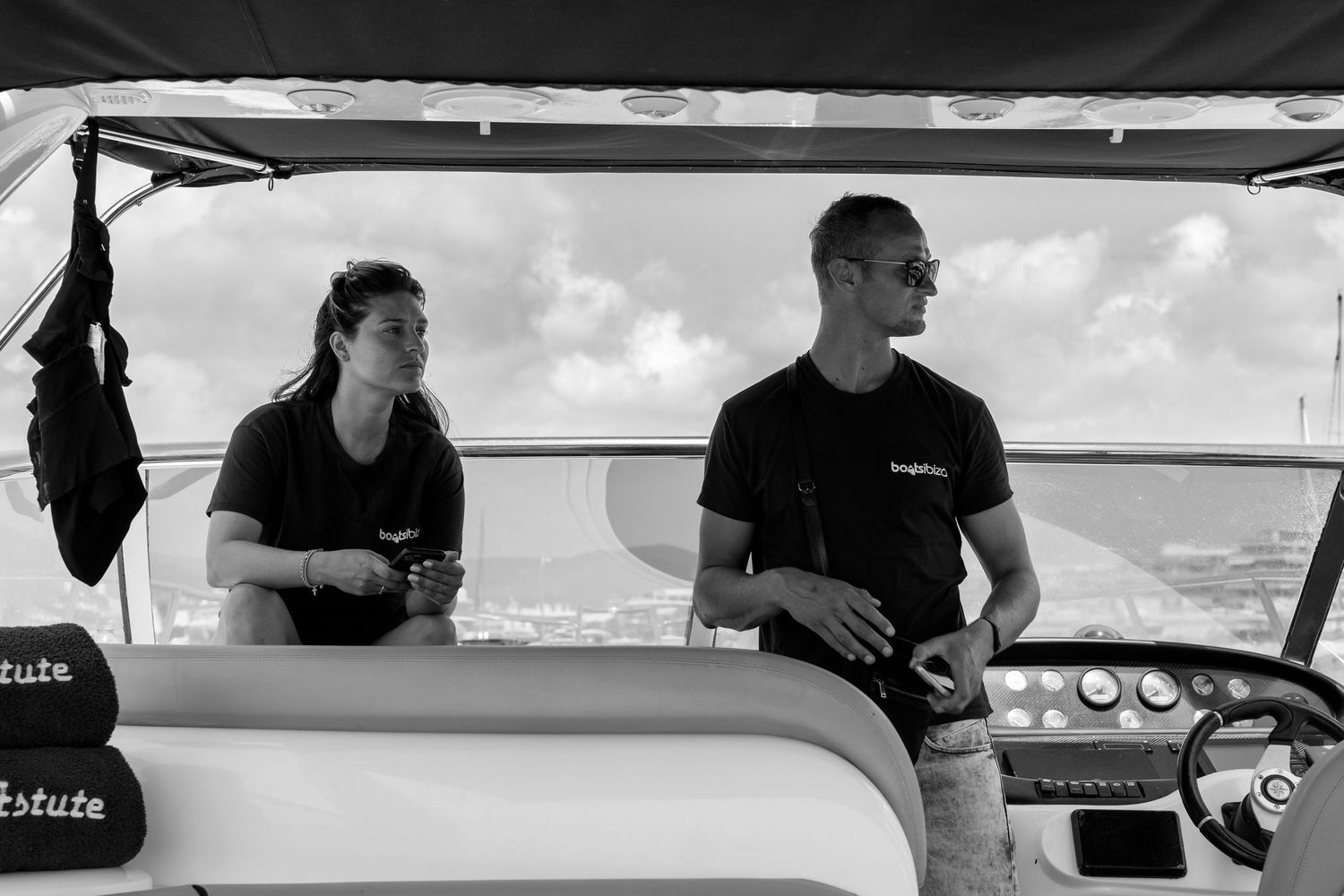 Two people in black shirts on a boat, man steering while woman sits beside him, ocean in background.