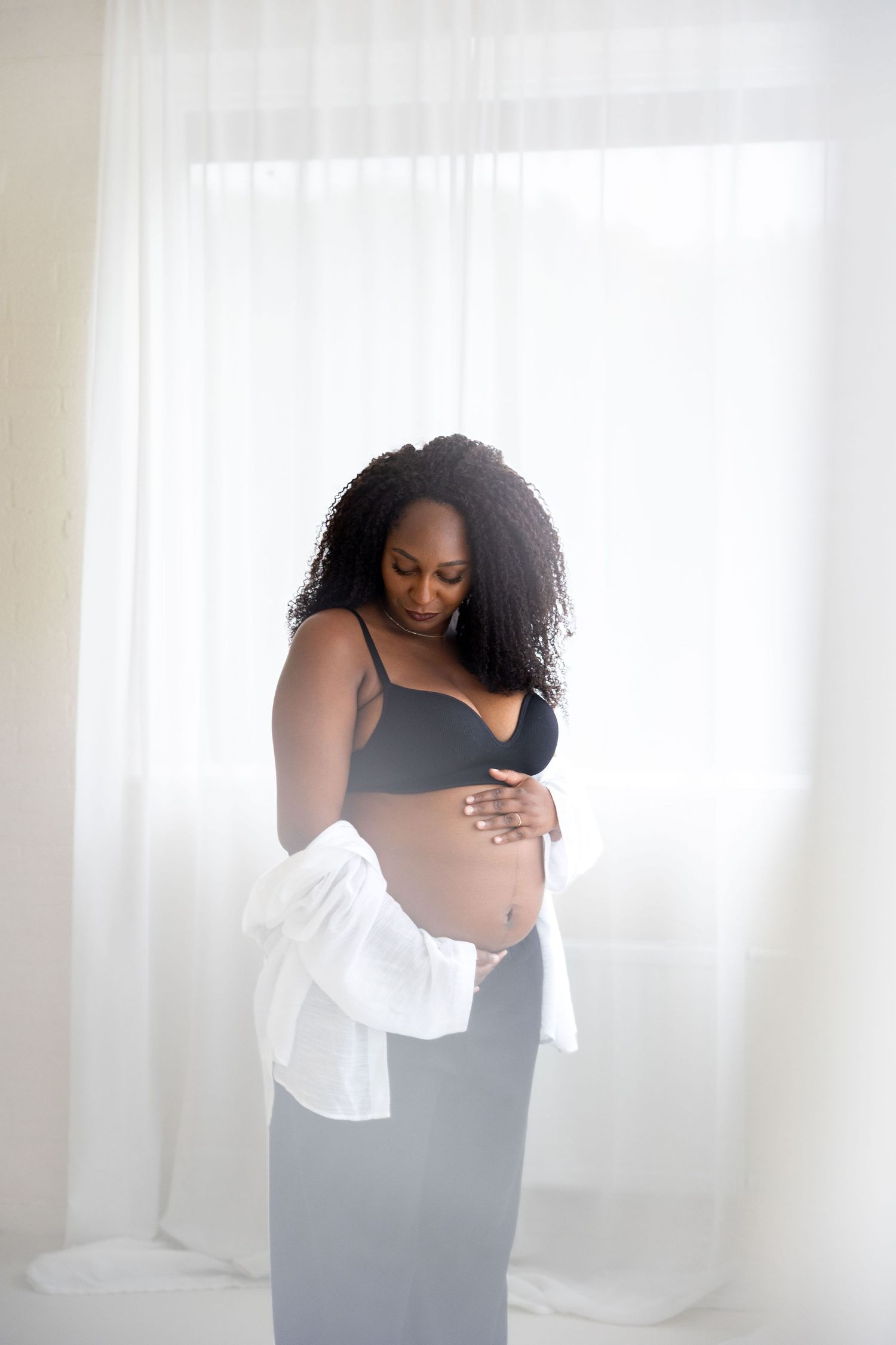 Pregnant Black woman in black bra and gray skirt holds baby bump, wearing open white shirt, standing by sheer curtains.