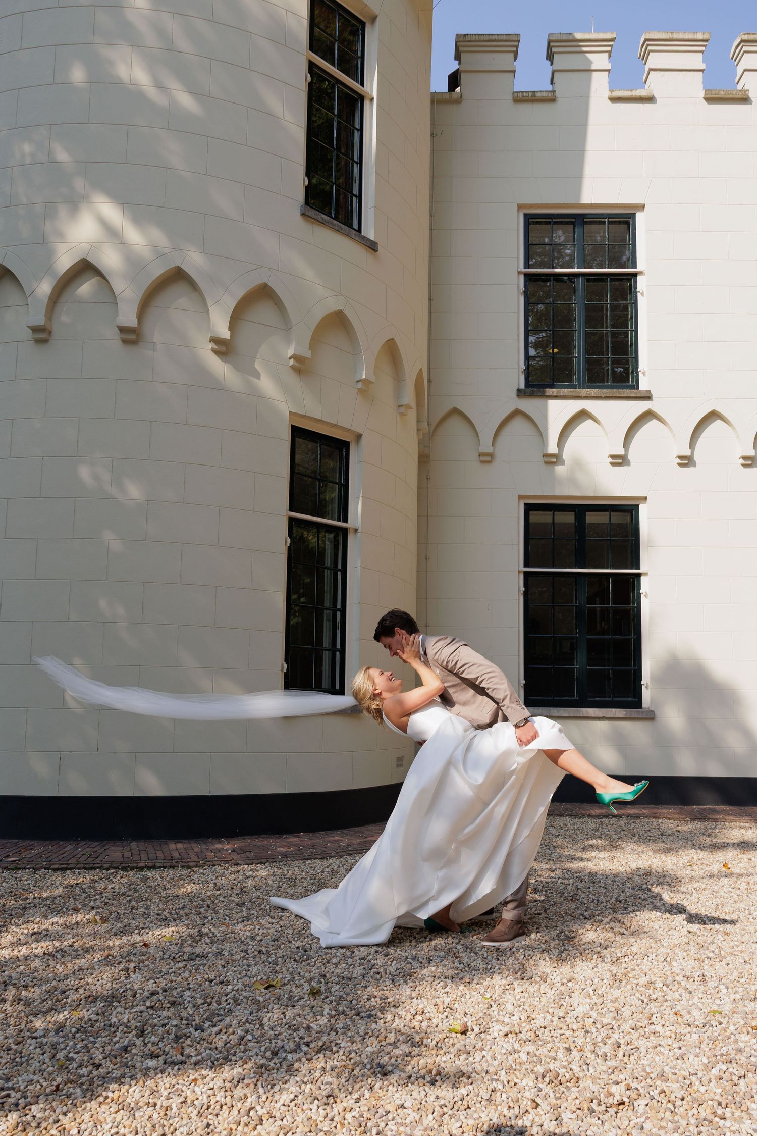 Bride and groom share a romantic dip kiss outside a white castle-style building on a sunny wedding day.
