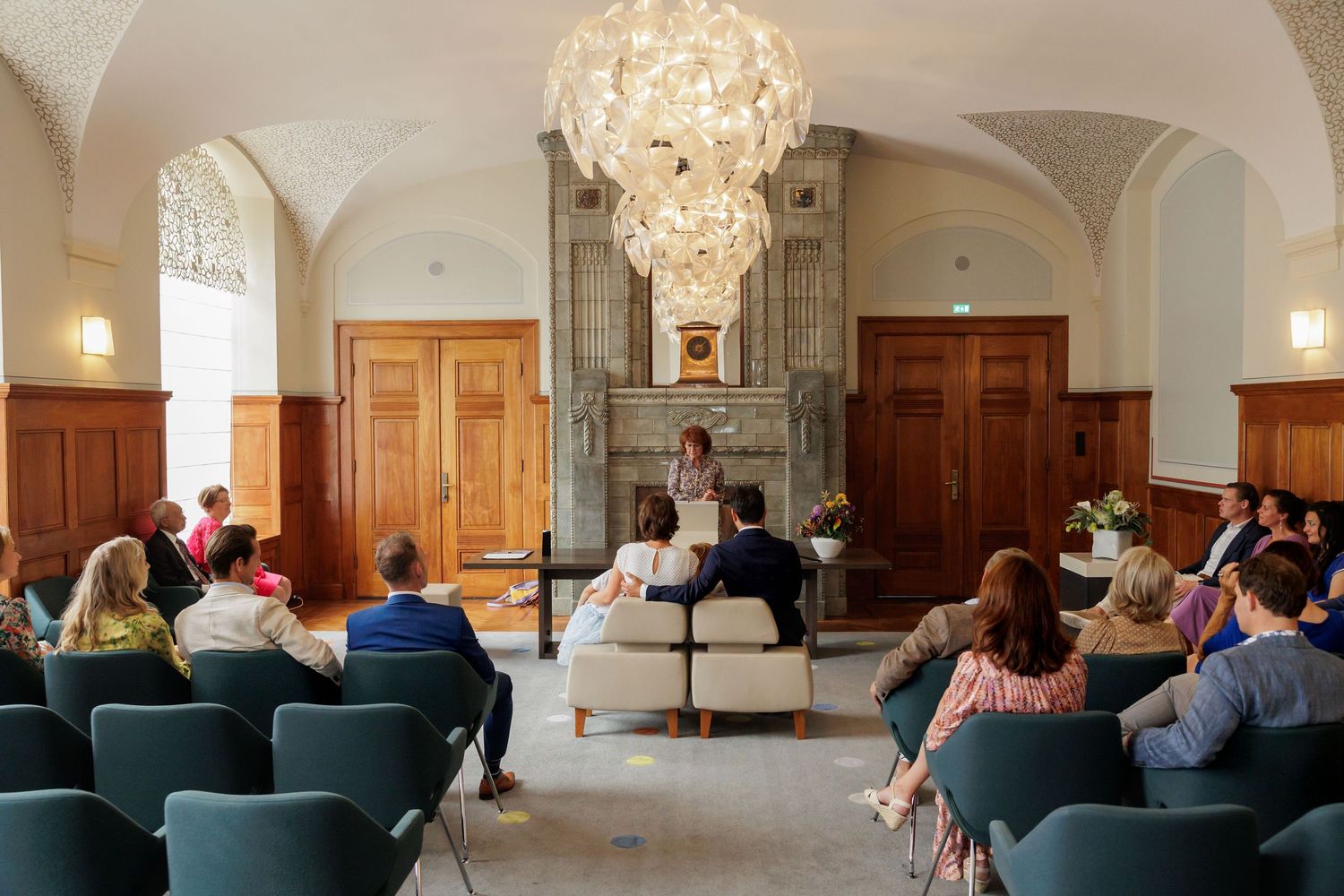 Elegant wedding ceremony in a grand hall with chandelier, guests seated in teal chairs facing a speaker near a stone fireplace.