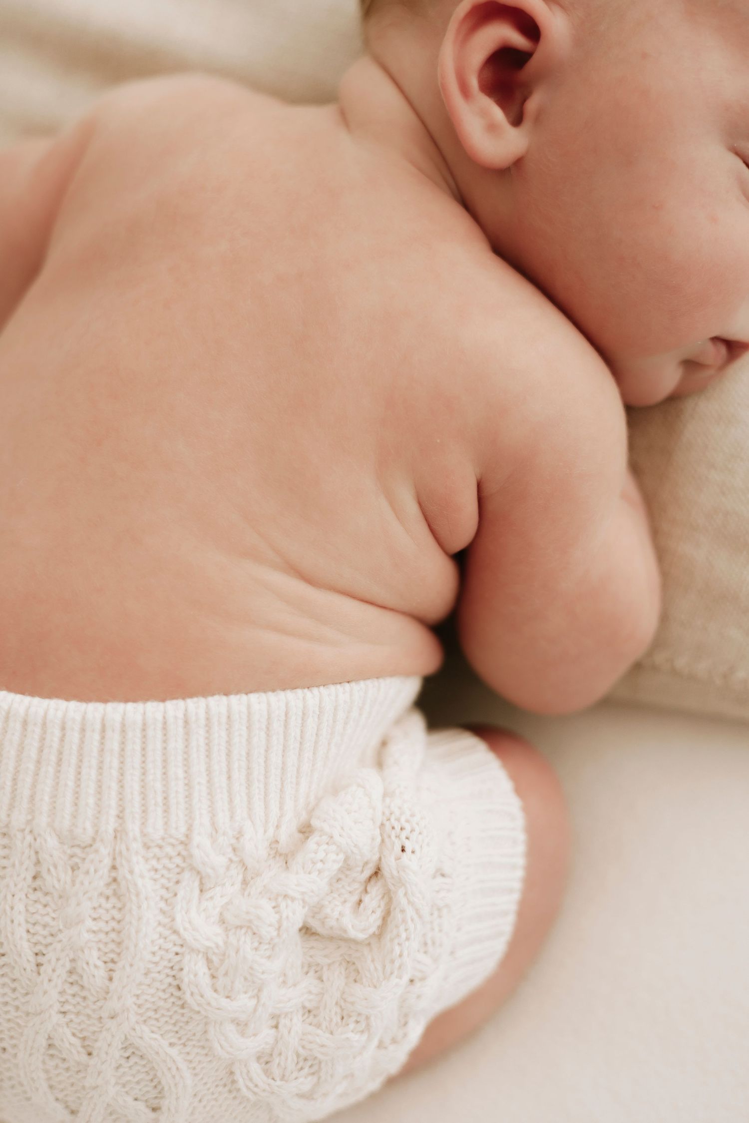 Sleeping newborn wearing white knitted clothing on cream colored bedding.