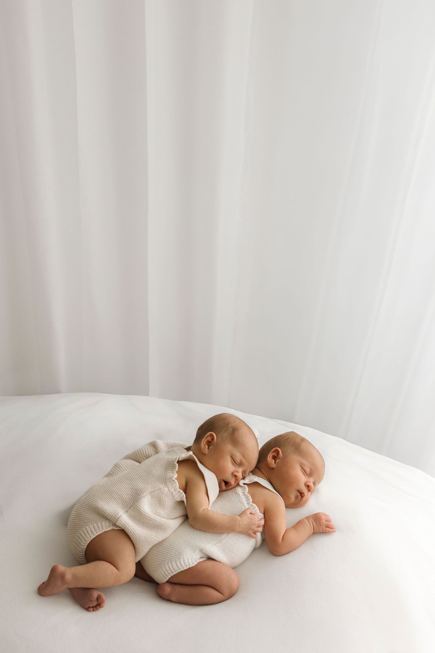 Two sleeping newborn twins in white knit rompers cuddle together on a soft white surface.