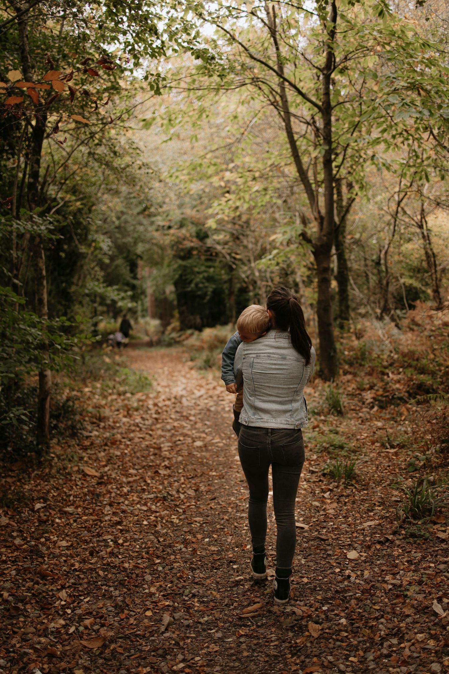 Person walking on a leaf-covered forest trail during autumn with trees lining the path.