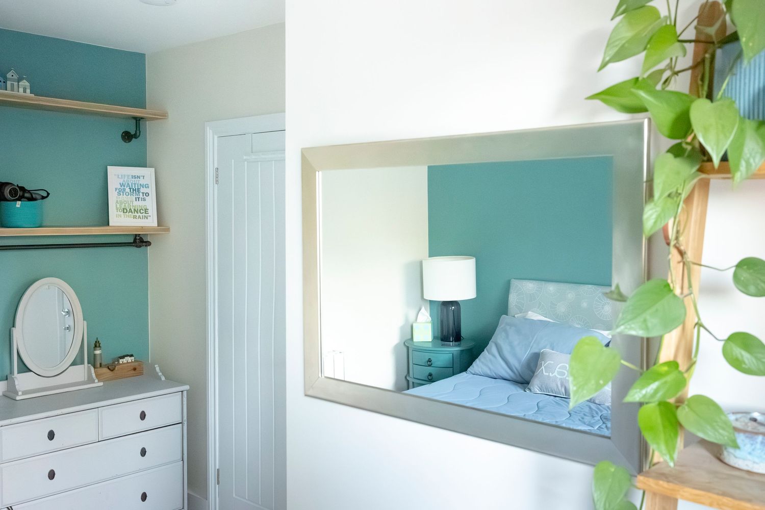 A bright bedroom with teal walls, a large mirror, white dresser, and lush green trailing pothos plant on a wooden shelf.