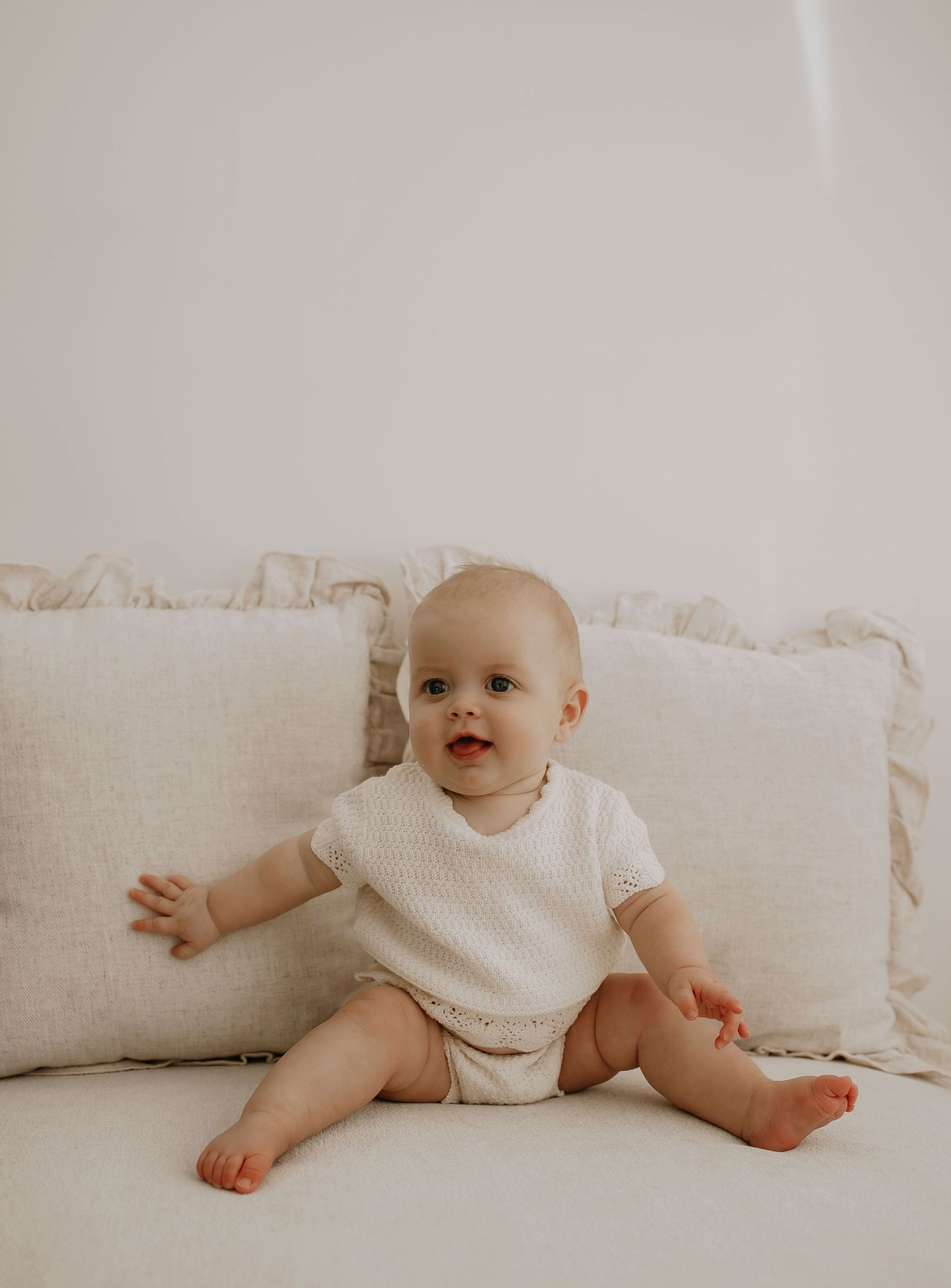 Baby in white outfit sitting on beige couch cushions.