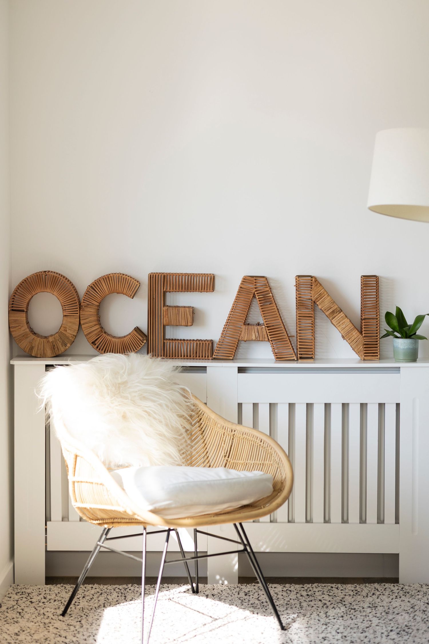 Coastal boho interior with rattan chair, fluffy pillow, and wicker OCEAN letters on white radiator cover.