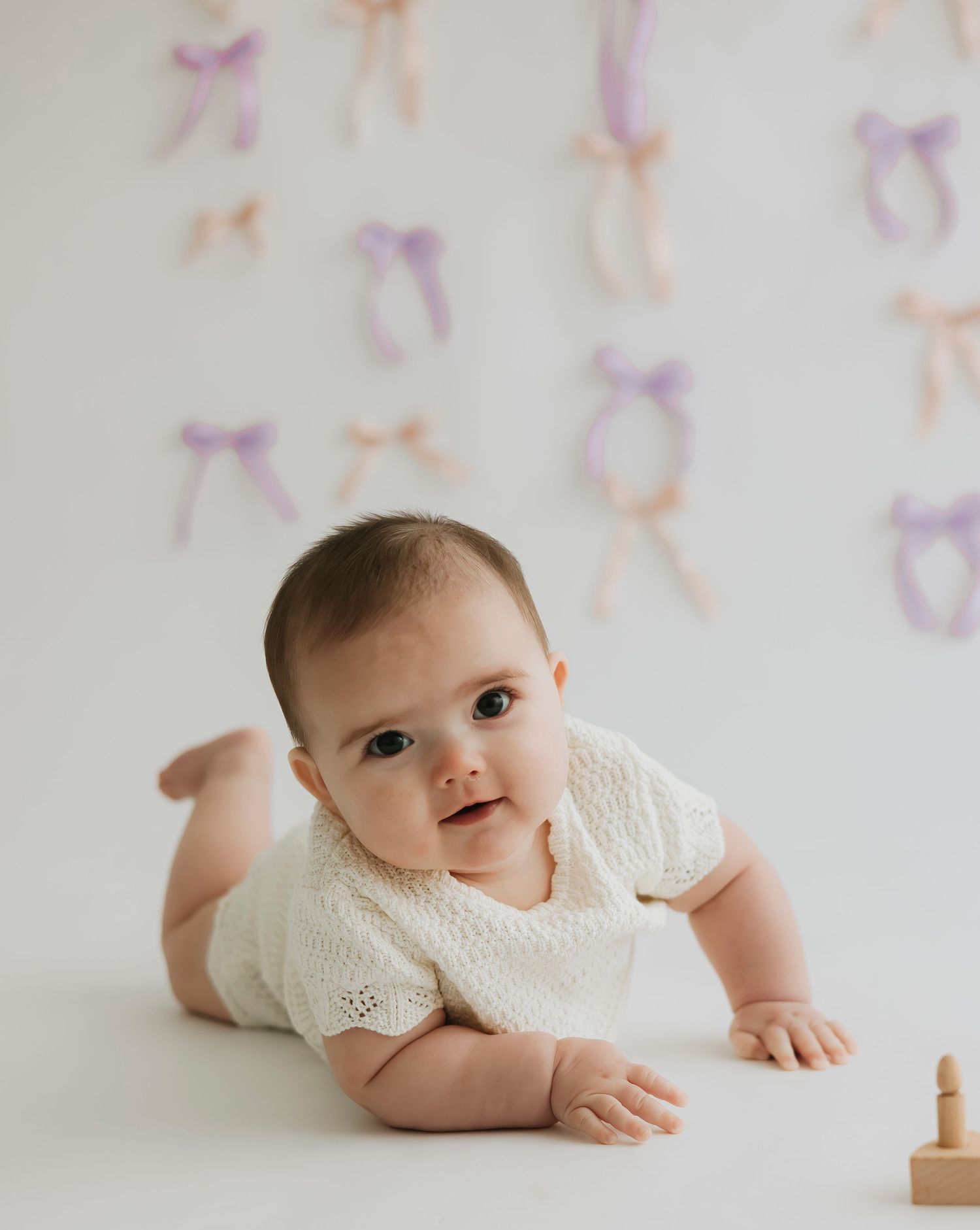 Baby in white romper lying on floor with bow pattern wallpaper.