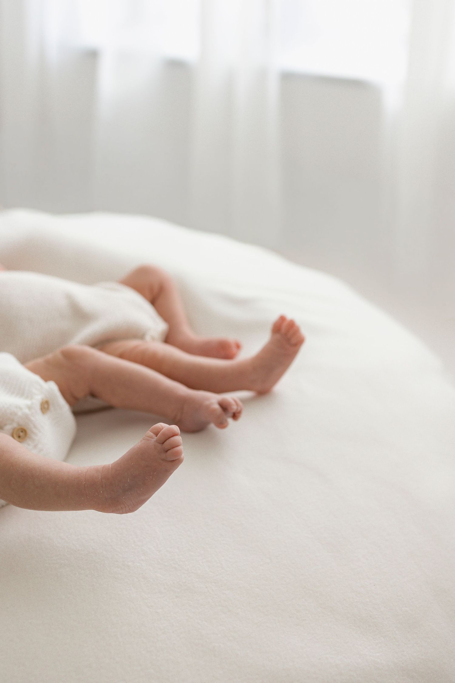 Close-up of two newborn babies' tiny feet and toes resting on a soft white blanket in a bright, airy room.