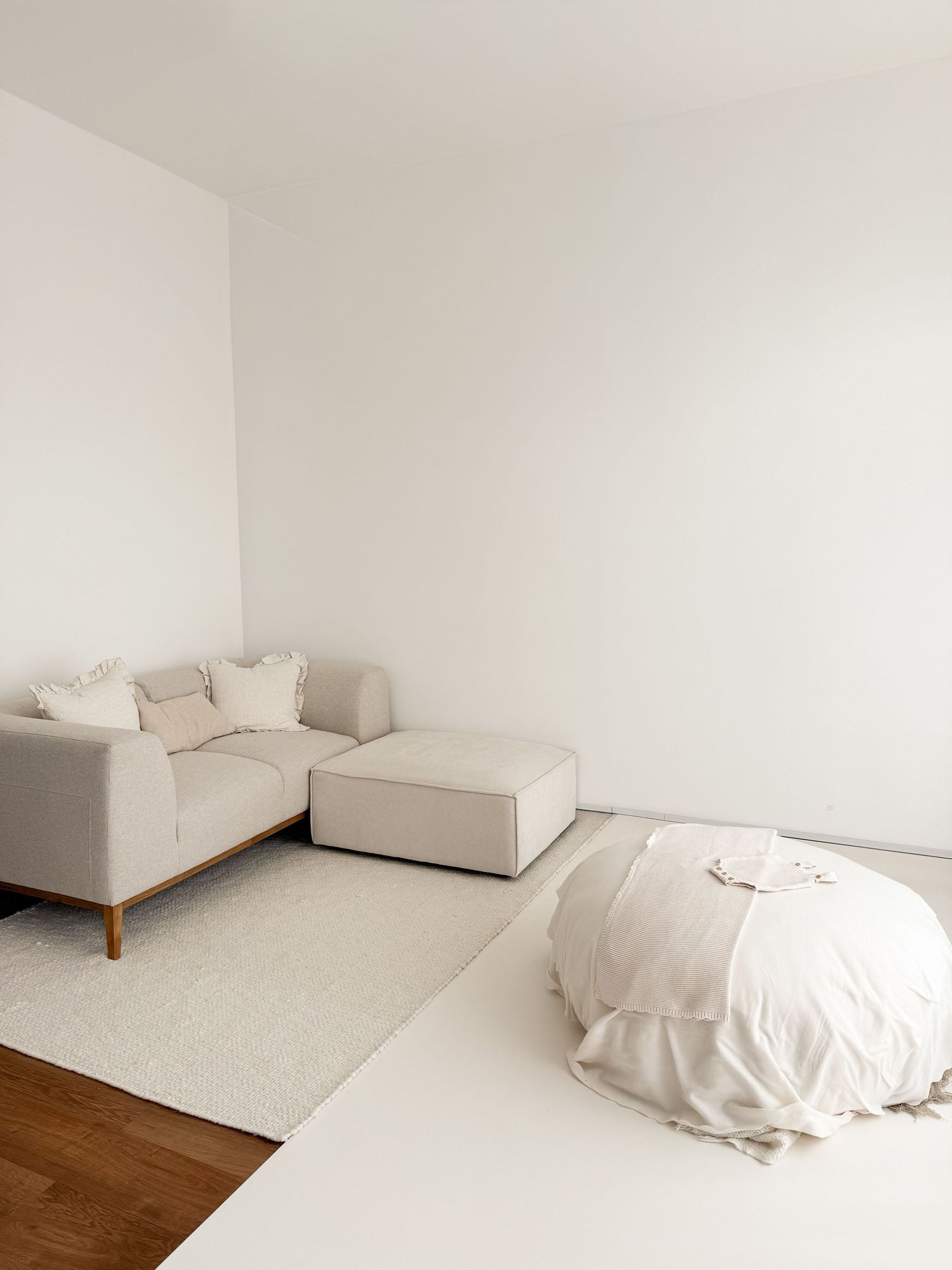 Minimalist white living room with a cream sectional sofa, ottoman, area rug, and floor cushion on light hardwood floors.