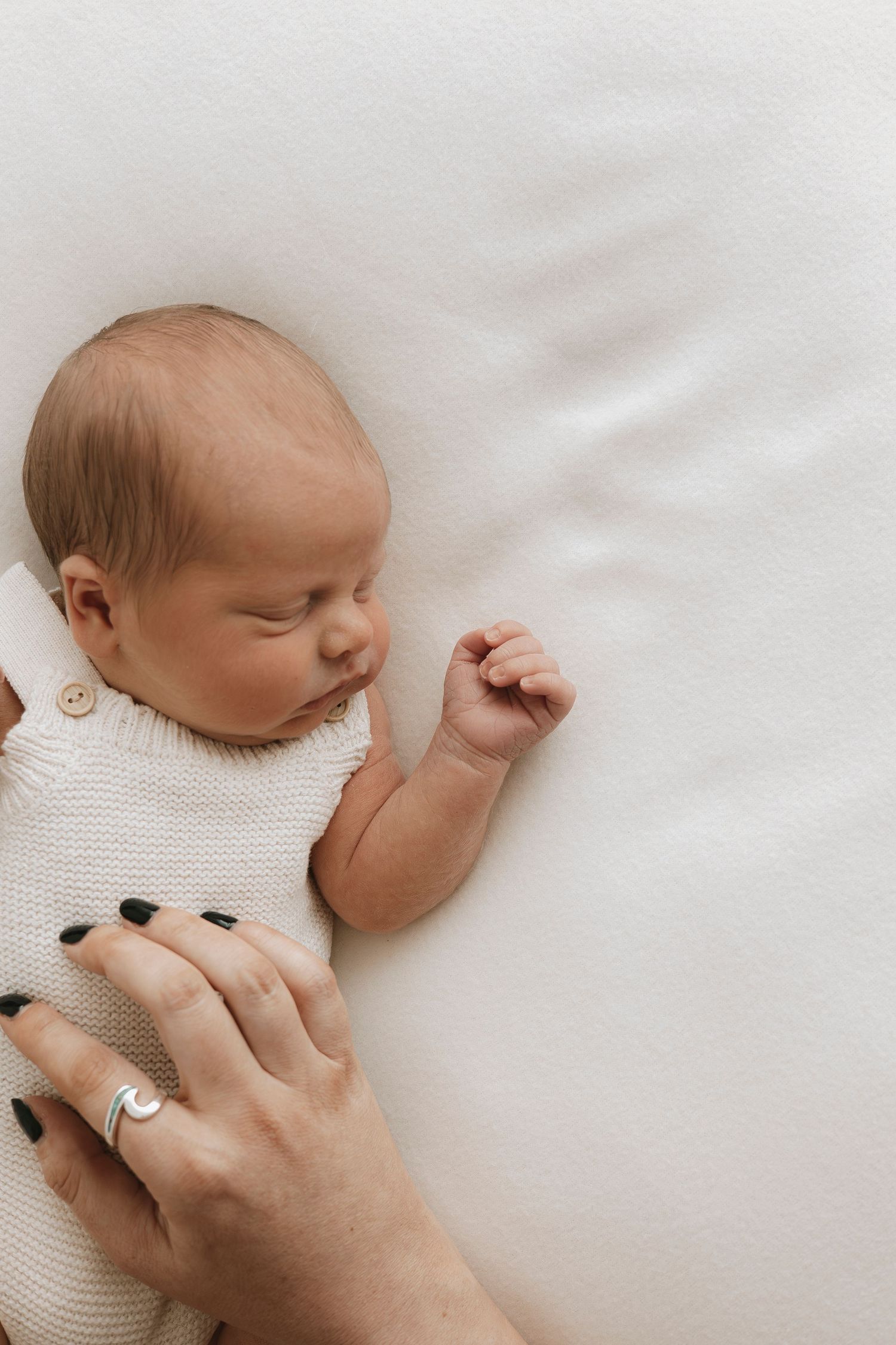 Sleeping newborn baby in cream knit romper lying on white blanket with mother's hand gently resting nearby.
