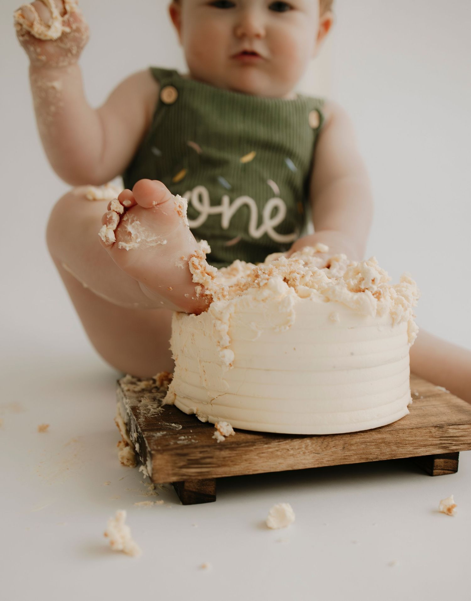 Small child in olive green outfit plays with white frosted cake on wooden board.