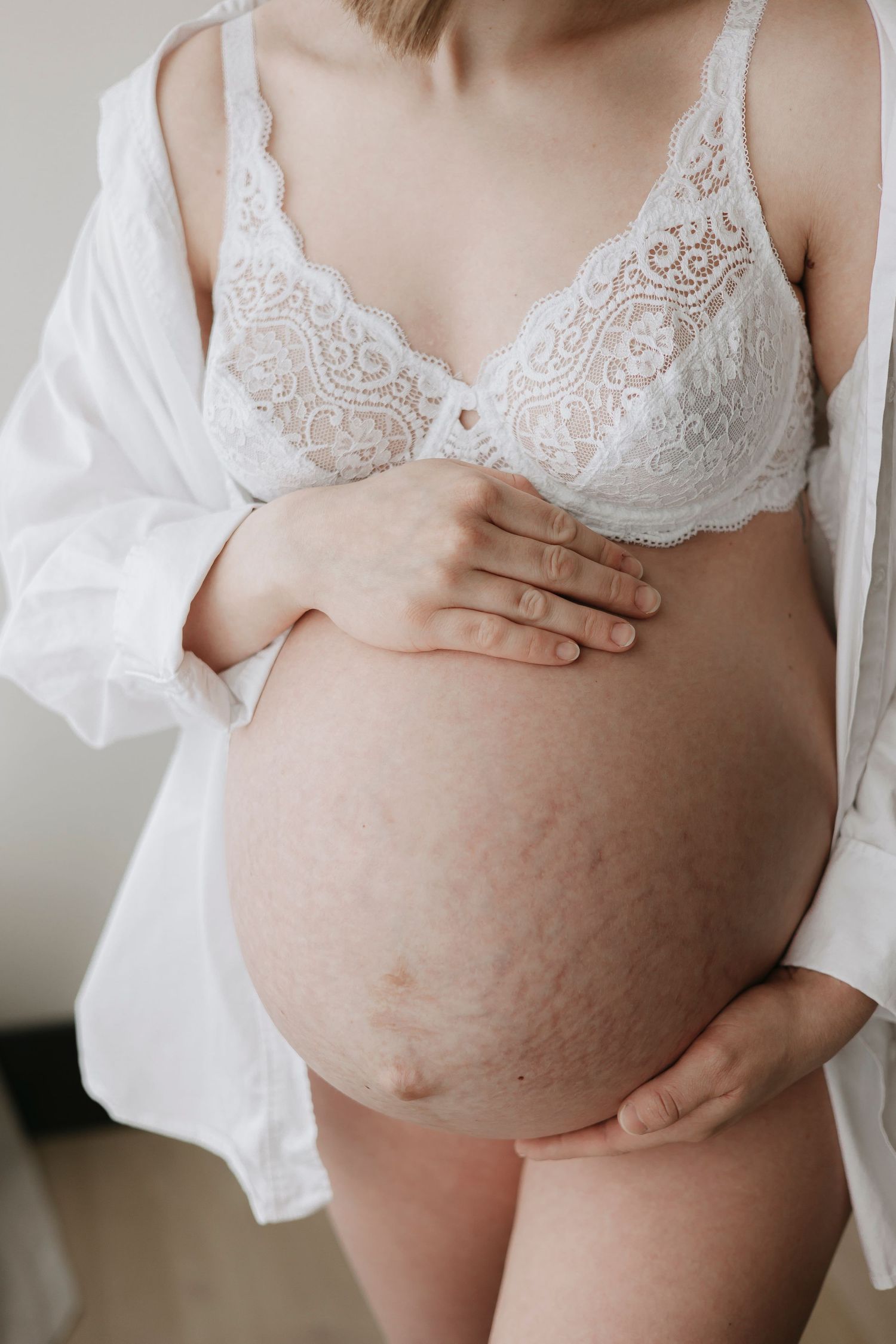 Close up view of a pregnant belly in white lace undergarments against light background.