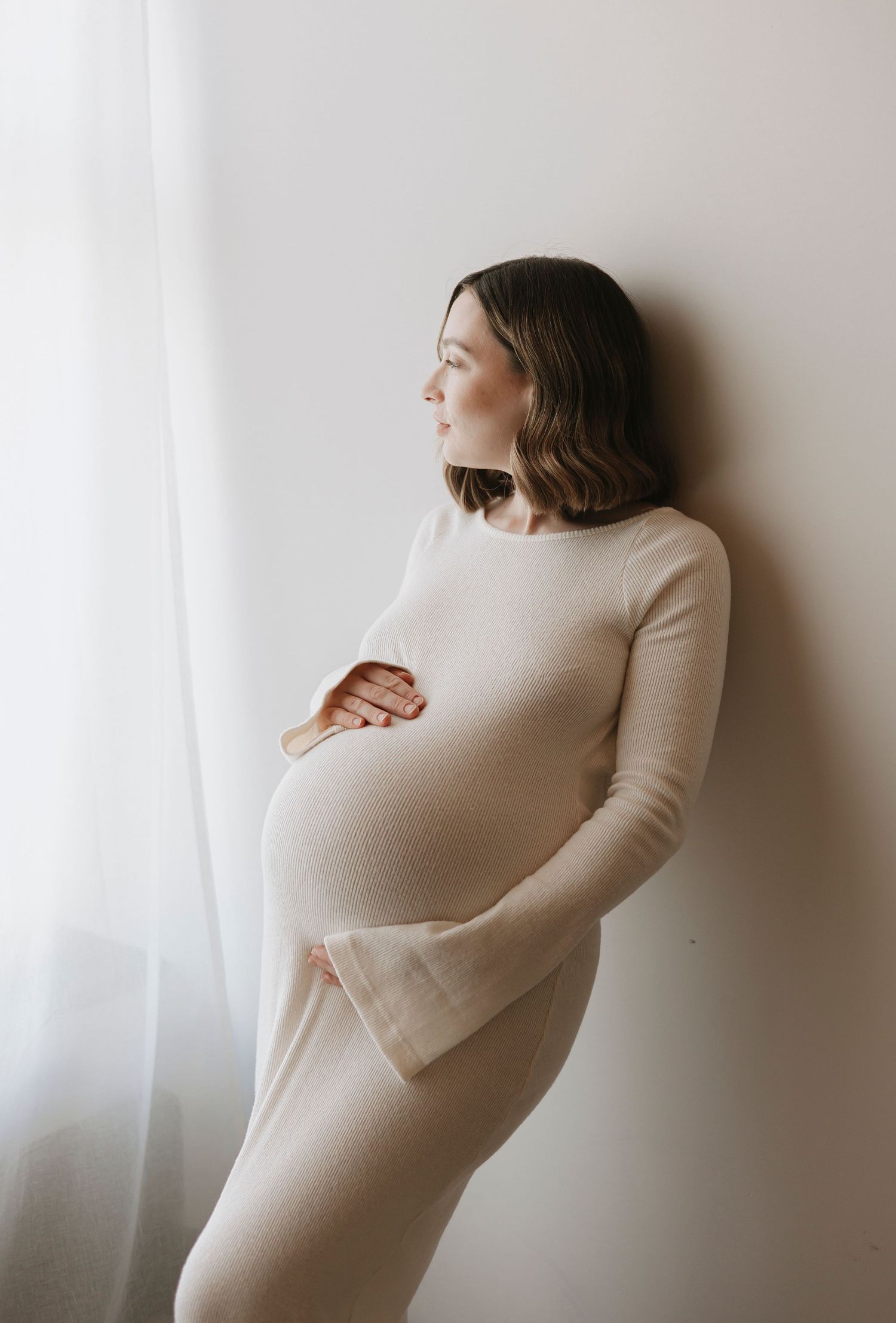 Elegant maternity portrait in beige sweater against white wall with natural window light.