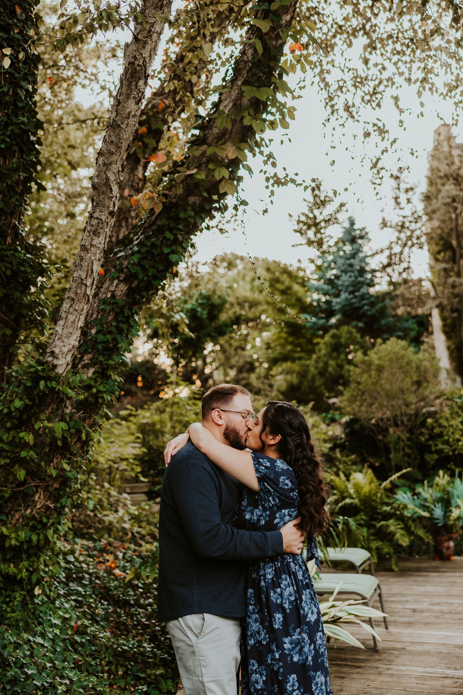 Romantic couple embracing in a lush garden setting with sunlit trees in the background.