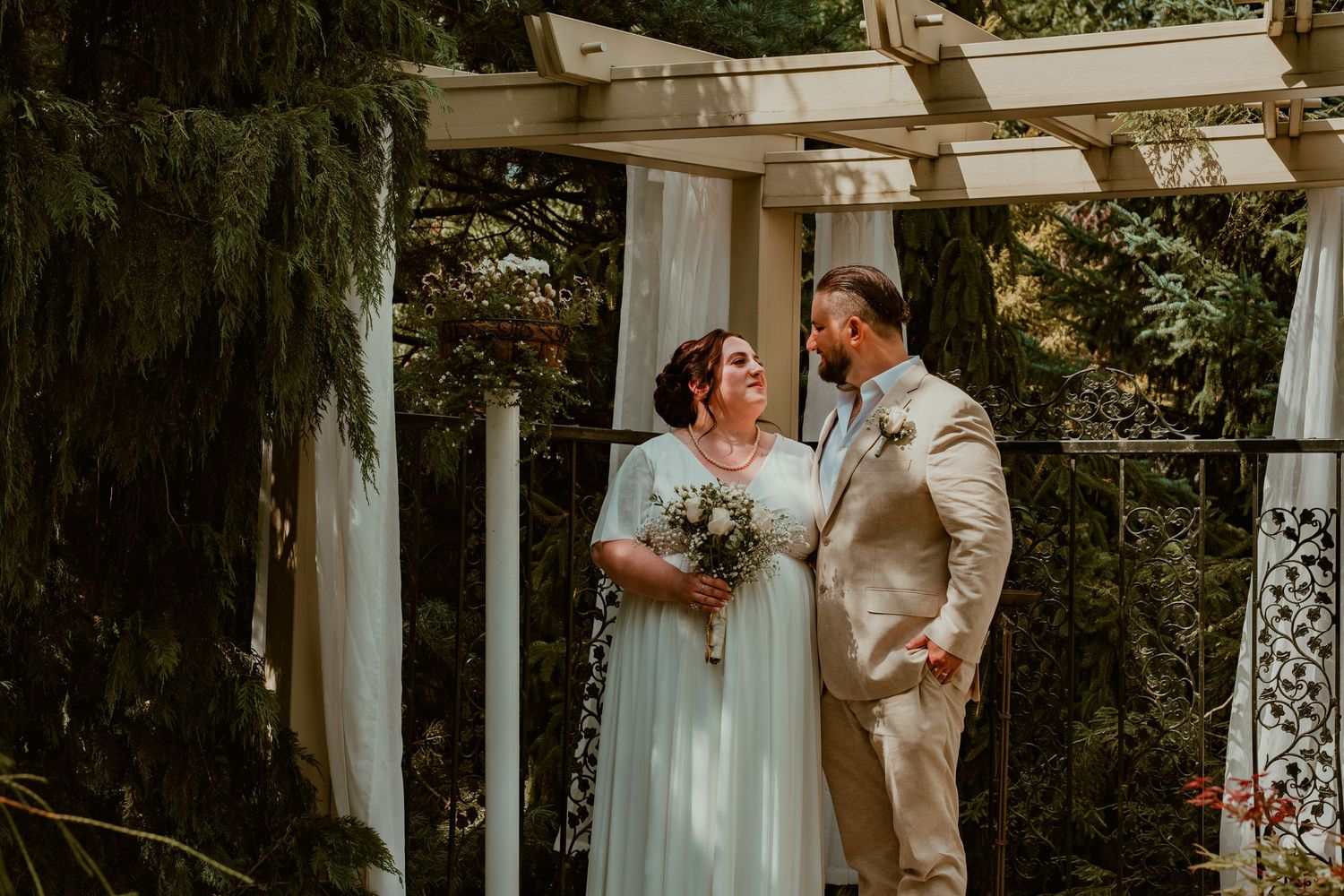 A romantic wedding ceremony takes place under a white pergola in a lush garden setting.