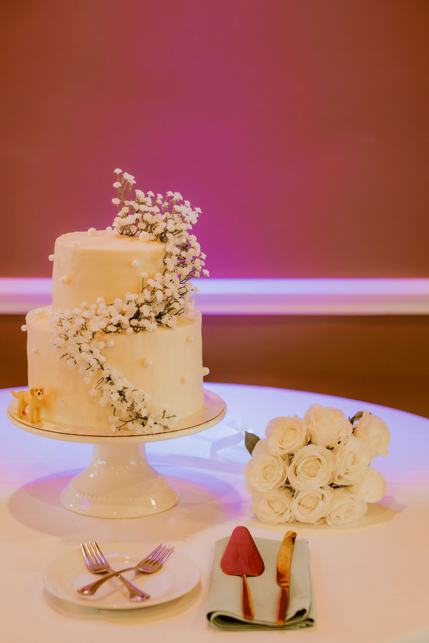 Elegant two-tier white wedding cake with cascading baby's breath decoration and pink lighting.
