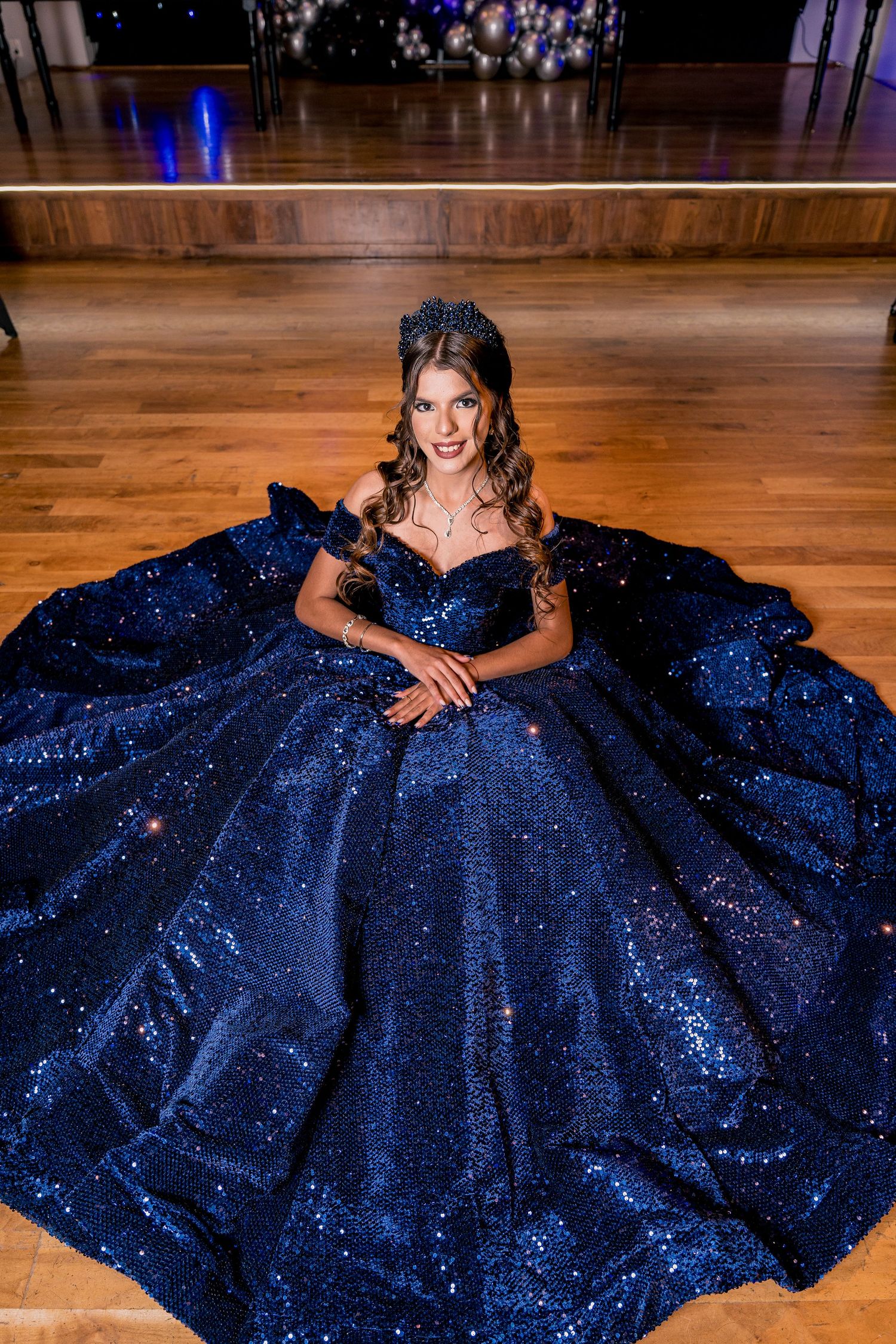 A sparkling navy blue ballgown with sequins spreads across a wooden dance floor under dramatic lighting.