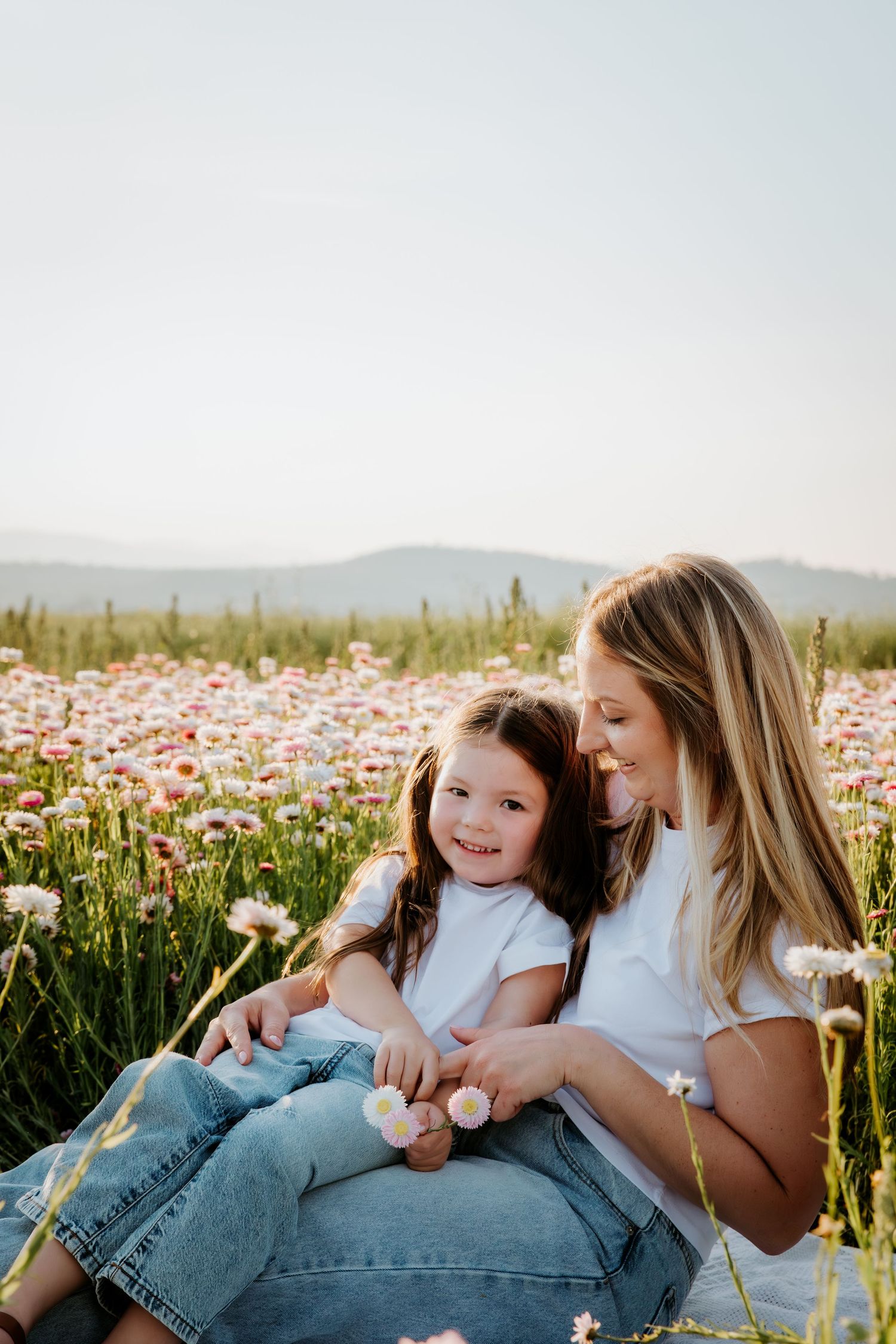A mother and young daughter sit together in a blooming flower field, both wearing white tees and jeans, smiling warmly.
