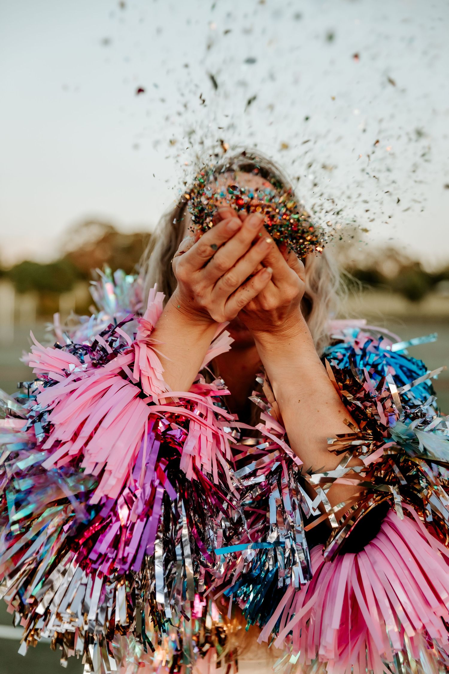 Colorful confetti and glitter explodes over hands covered in festive pink and blue metallic fringe decorations.