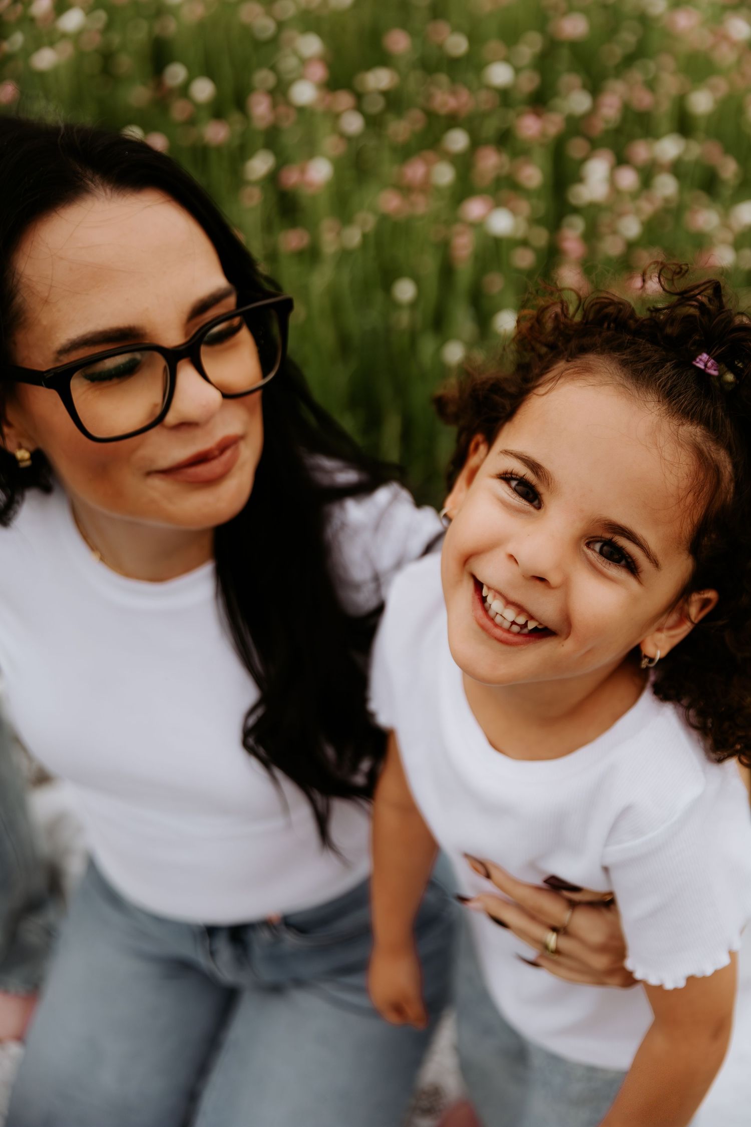 A woman with glasses and a young girl in matching white shirts smile together in a flower field.