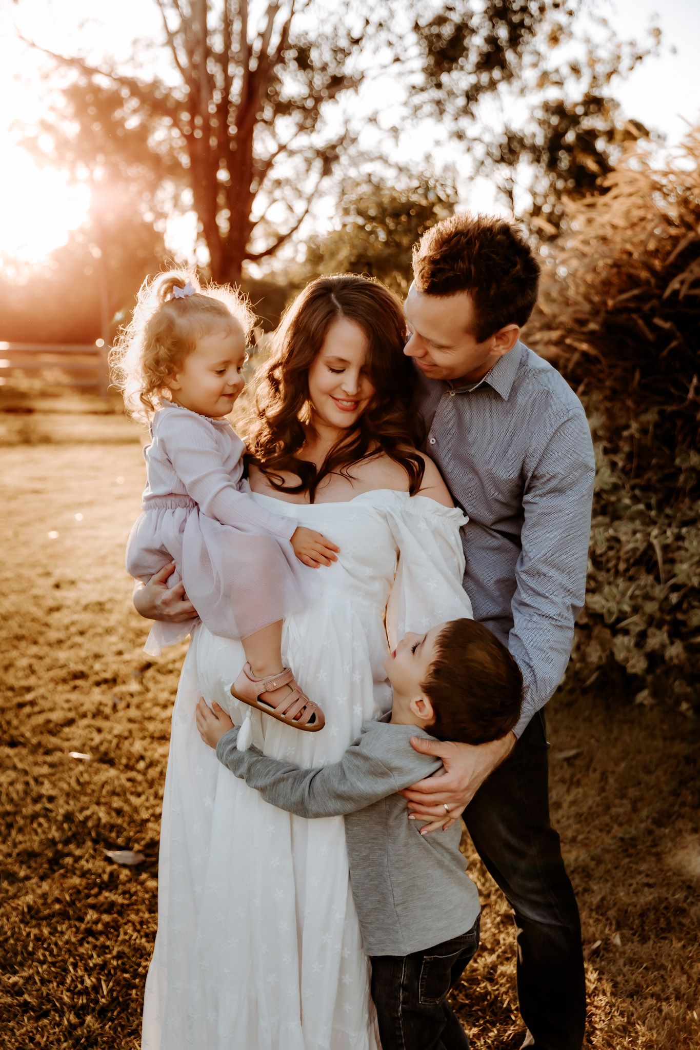 Family photoshoot at sunset in a field with warm golden light filtering through trees.