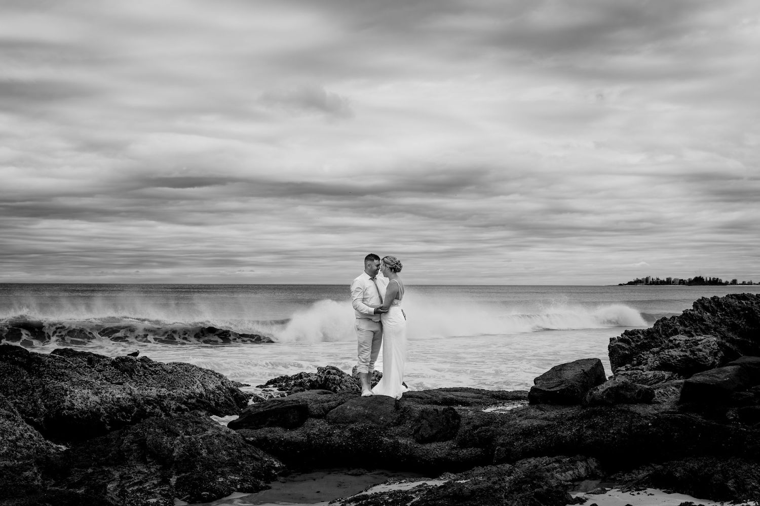 Dramatic black and white coastal wedding photos with cloudy sky and rocky shoreline.