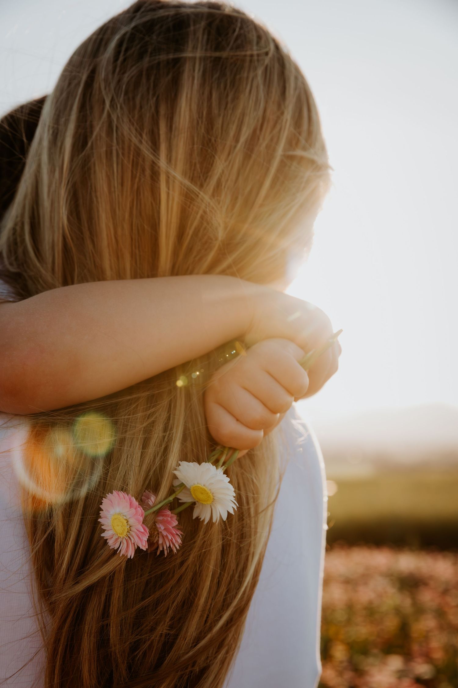A girl with long blonde hair holds pink and white daisies, backlit by golden sunlight in a flower field.