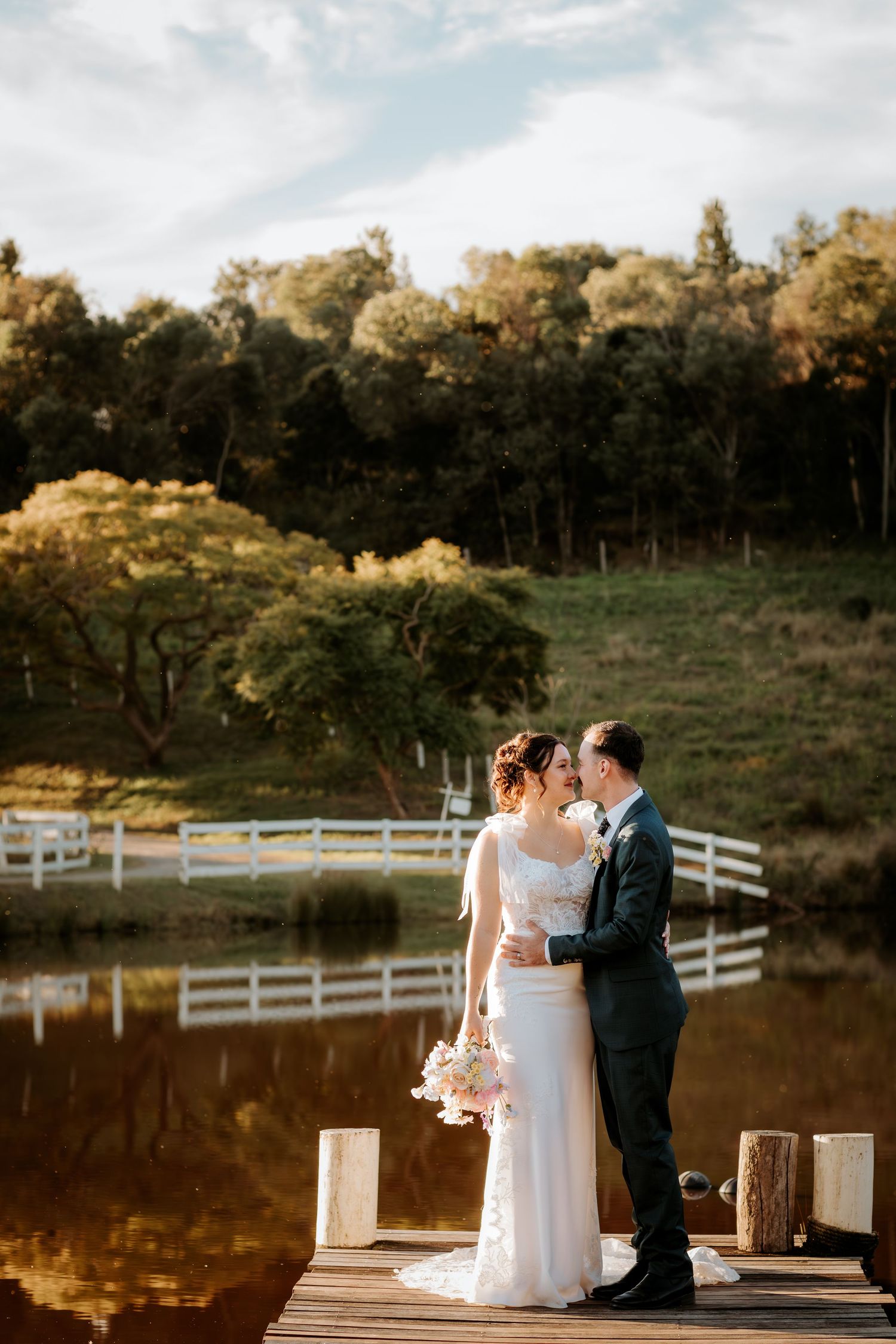 Newlyweds standing on wooden dock by lake with white fencing in background at sunset.
