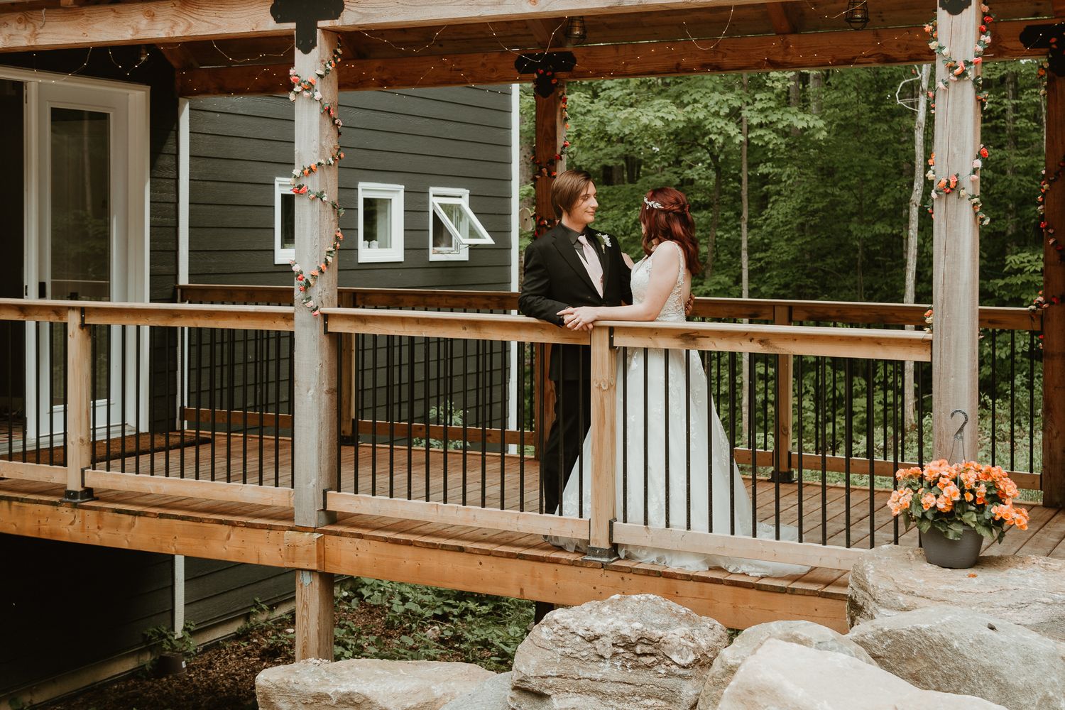 A wooden deck with white railing posts extends from a green house surrounded by lush forest scenery.