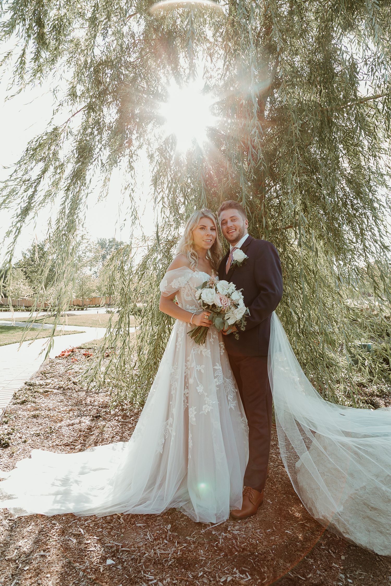 A couple poses under a weeping willow tree on their wedding day with sunlight streaming through the branches.
