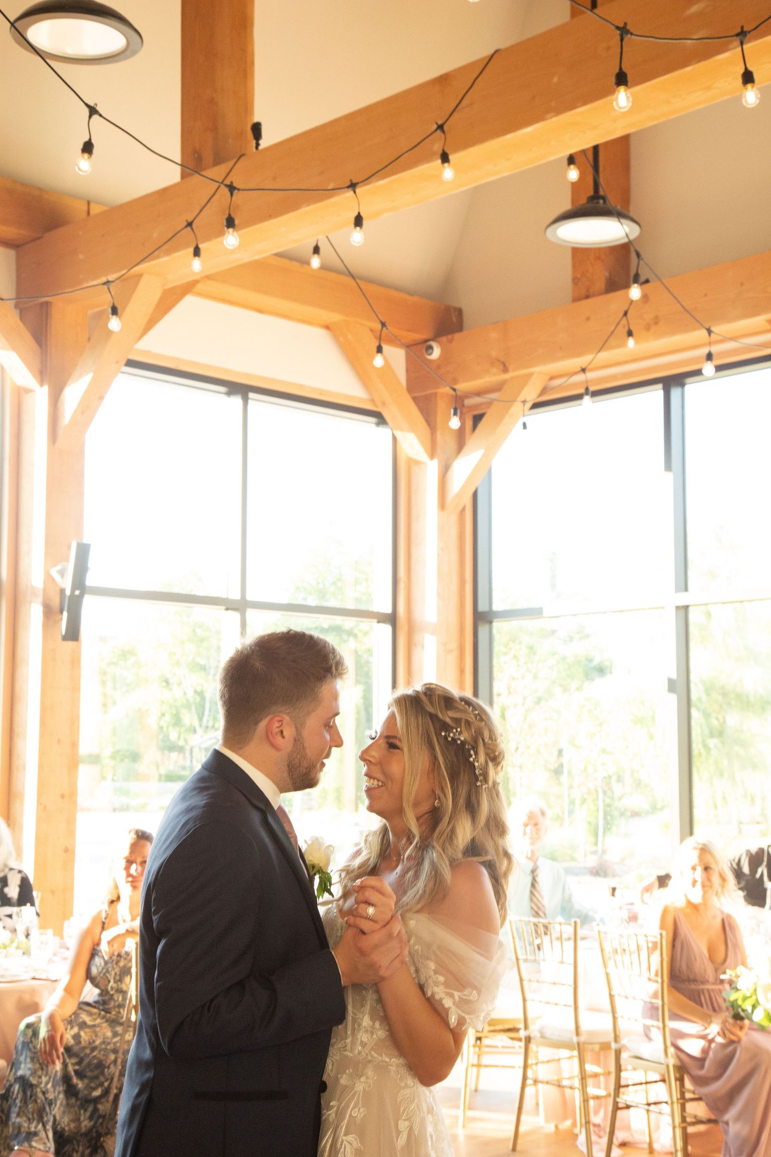 Couple shares first dance at wedding reception with string lights and large windows in warm-lit venue.